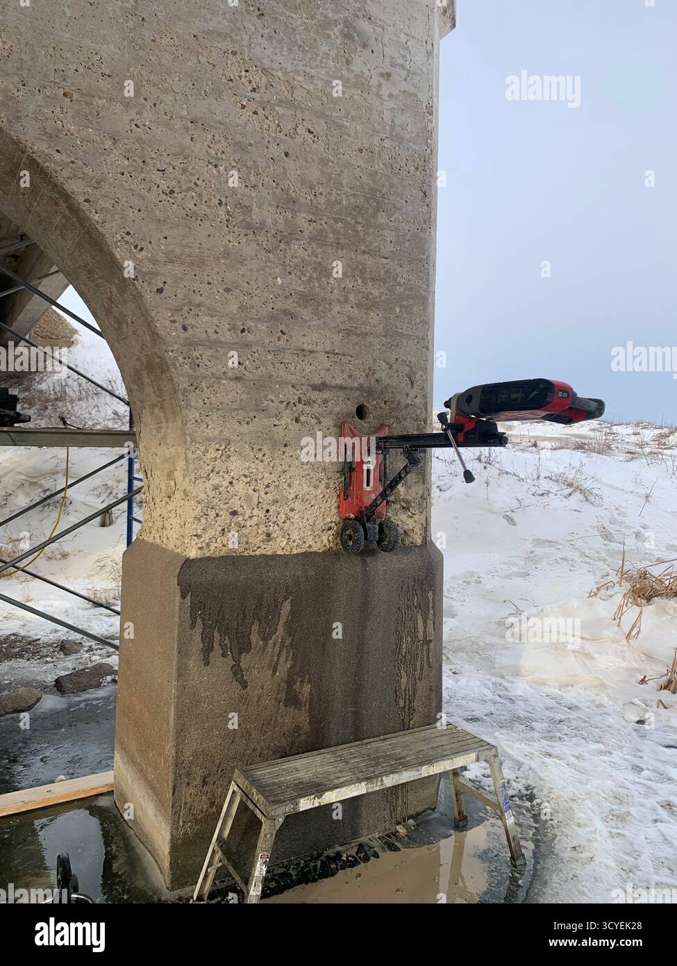 Reparatur von Winterbrücken: Kernbohrer montiert auf verschneiten Betonpier über eiskaltem Fluss, kalte Bauszene. - Smartphone-aufgenommenes Stockfoto