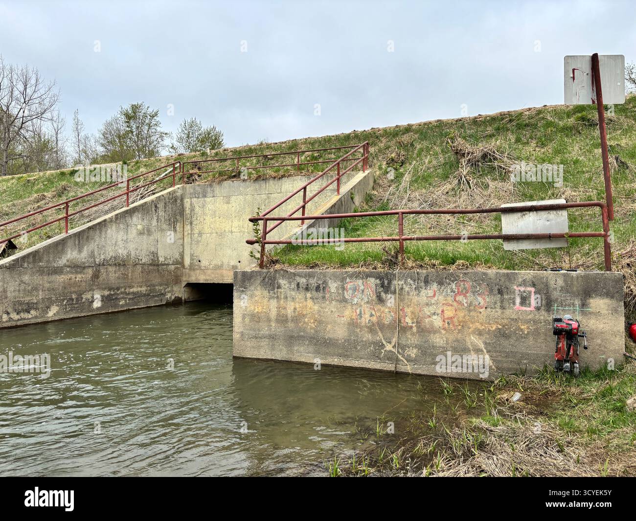 Betonabflusskanal mit rotem Geländer und Wasserfluss darunter, Teil der Regenwasser- oder Bewässerungsinfrastruktur in einem ländlichen Gebiet. - Smartphone-aufgenommenes Stockfoto