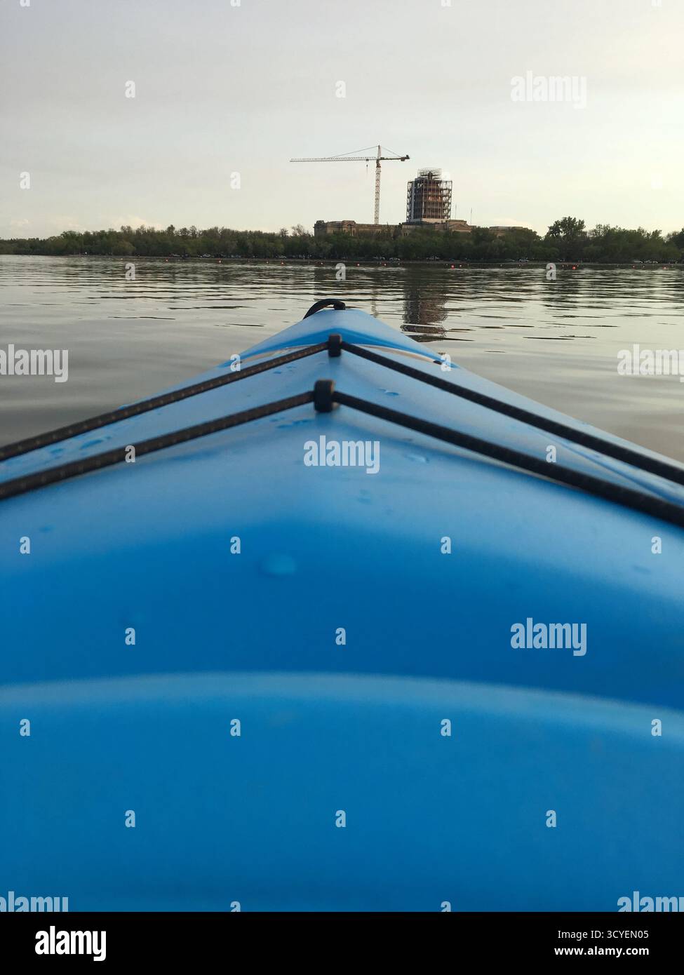Kajak gleitet auf ruhigem Wasser bei Sonnenuntergang, mit einem Kran und einer Silhouette vor einem goldenen Himmel, die Natur und Stadt verbindet. Stockfoto