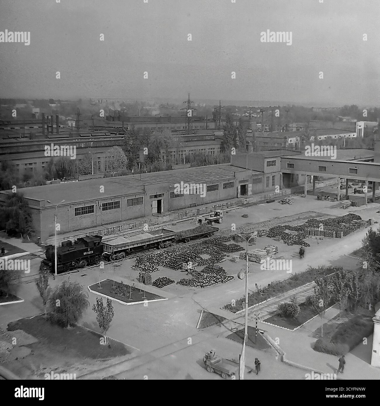 Dieses archivierte Schwarzweißfoto aus den 1970er Jahren zeigt einen Hochwinkel-Panoramablick auf die Slavjansk Armature-Isolator Plant (AIZ) in Sloviansk, UdSSR. Der riesige Industriehof dient als Freiluftlager, gefüllt mit massiven Häufern fertiger Isolatoren. Die Bilder dokumentieren die Logistik der Fabrik und zeigen eine kleine Rangierlokomotive (Nr. 8223) auf dem Gleisanschluss sowie schwere Gabelstapler, die Güter transportieren. Das ist ein Dokument sowjetischen industriellen Ausmaßes Stockfoto