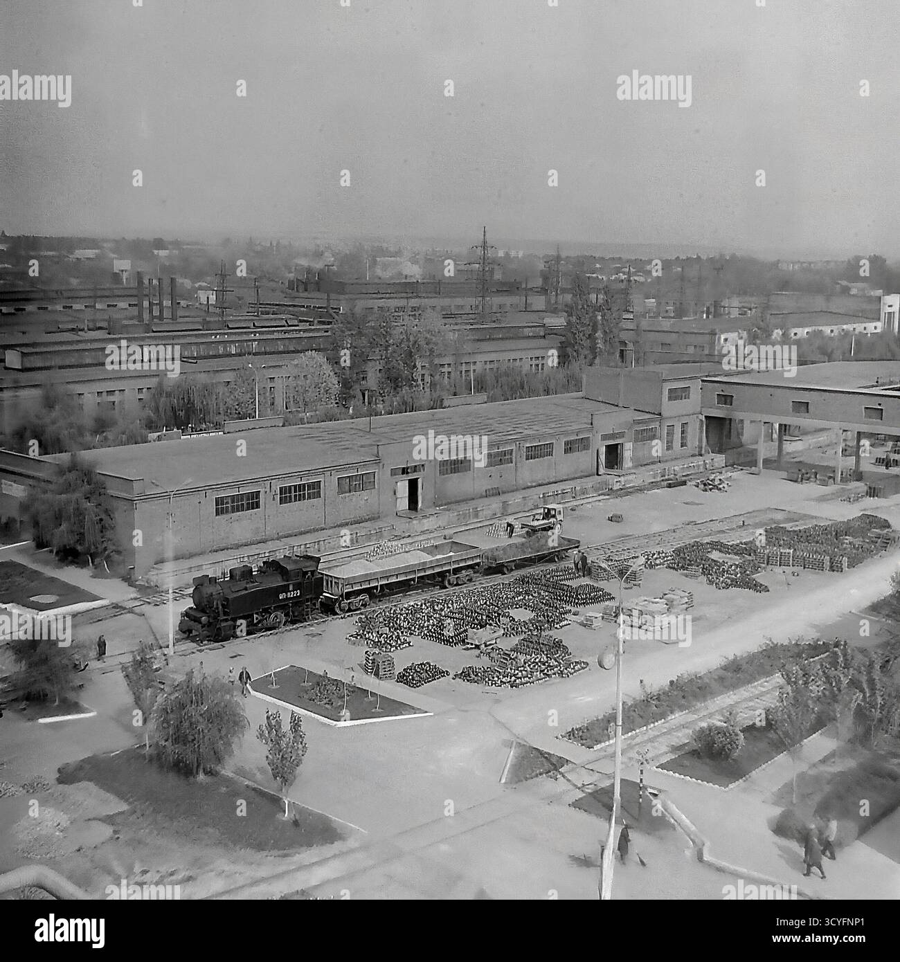 Dieses archivierte Schwarzweißfoto aus den 1970er Jahren zeigt einen Hochwinkel-Panoramablick auf die Slavjansk Armature-Isolator Plant (AIZ) in Sloviansk, UdSSR. Der riesige Industriehof dient als Freiluftlager, gefüllt mit massiven Häufern fertiger Isolatoren. Die Bilder dokumentieren die Logistik der Fabrik und zeigen eine kleine Rangierlokomotive (Nr. 8223) auf dem Gleisanschluss sowie schwere Gabelstapler, die Güter transportieren. Das ist ein Dokument sowjetischen industriellen Ausmaßes Stockfoto