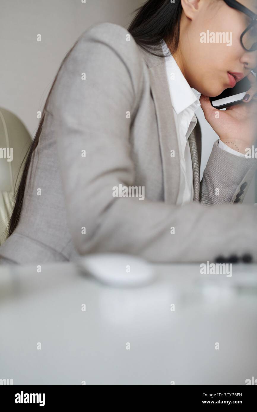 Junge asiatische Geschäftsfrau in elegantem Anzug und Brille, die auf dem Smartphone spricht, während sie am Bürotisch sitzt, mit flachem Blickwinkel Stockfoto