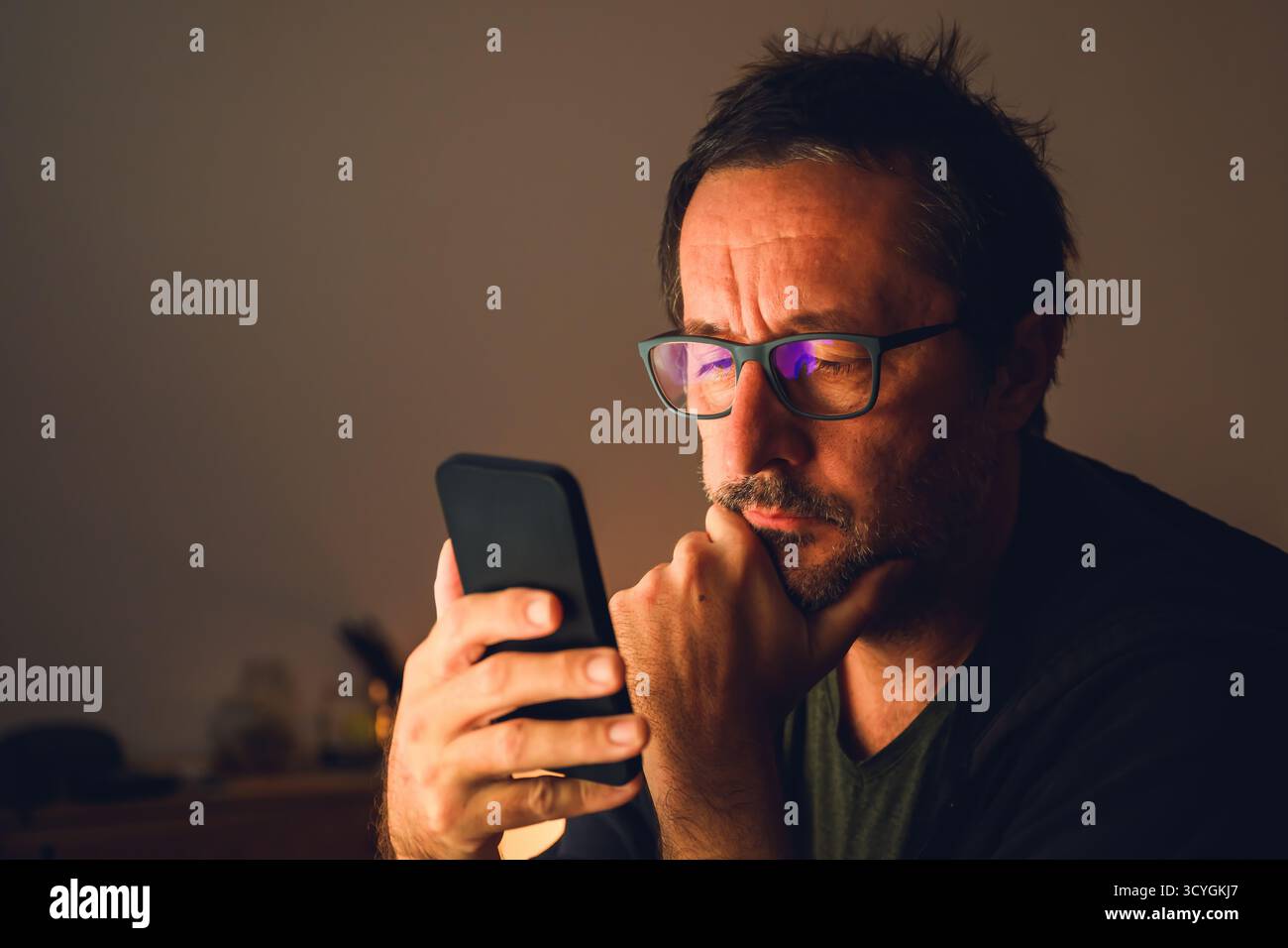Mann mittleren Alters mit Smartphone in schwach beleuchtetem Raum, fokussiert auf den Bildschirm mit Reflexion von blauem Licht in der Brille. Selektiver Fokus. Stockfoto