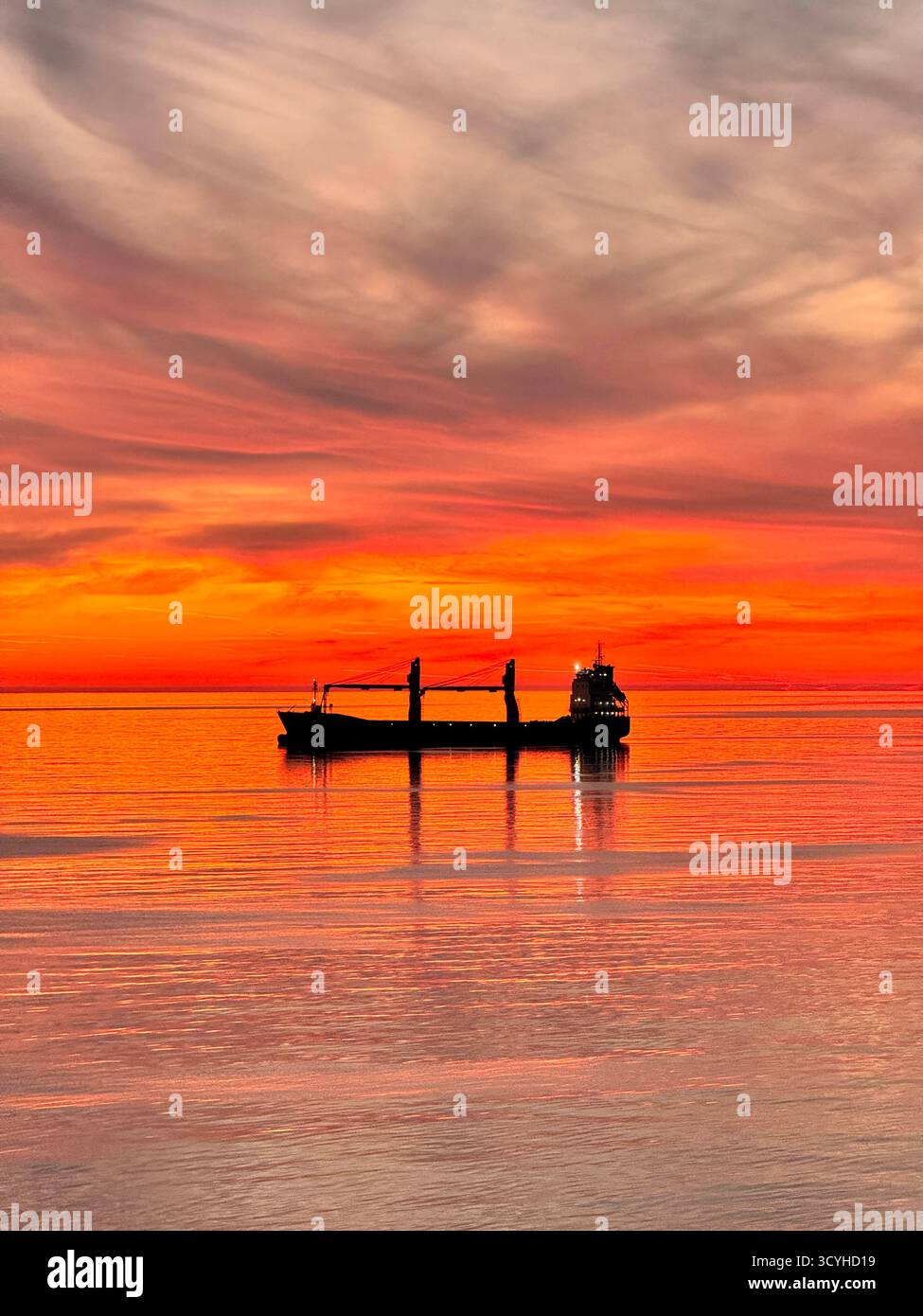 Ein Frachtschiff, das über ruhiges Wasser gleitet, während der Himmel in orange und goldenen Sonnenuntergangstönen brennt. Friedliche und filmische Meereslandschaft. Stockfoto