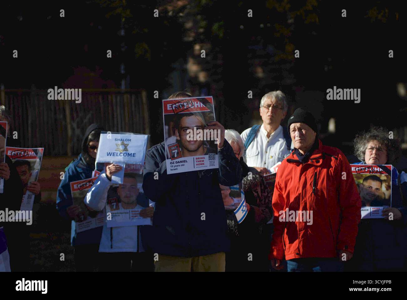 Flensburg, Schleswig-Holstein Gedenkstunde für israelische Geiseln auf dem Flensburger Südermarkt. Hier: Eine Person hält ein Plakat mit dem Porträt und Namen des Geisels Oz Daniel sowie den typischen Schriftzügen ENOUGH, OKT 7 und BRING SIE NACH HAUSE hoch. Links daneben ist ein weiteres Schild mit der Aufschrift Shalom Willkommen zu erkennen. Im Hintergrund und am rechten Bildrand stehen weitere Menschen, die ebenfalls Geisel-Plakate halten. Die Gruppe steht eng beieinander, die Gesichter der Teilnehmenden wirken ernst und anteilnehmend. Aufnahme vom 19.10.2025, Flensburg *** Flensburg, Schleswig Ho Stockfoto