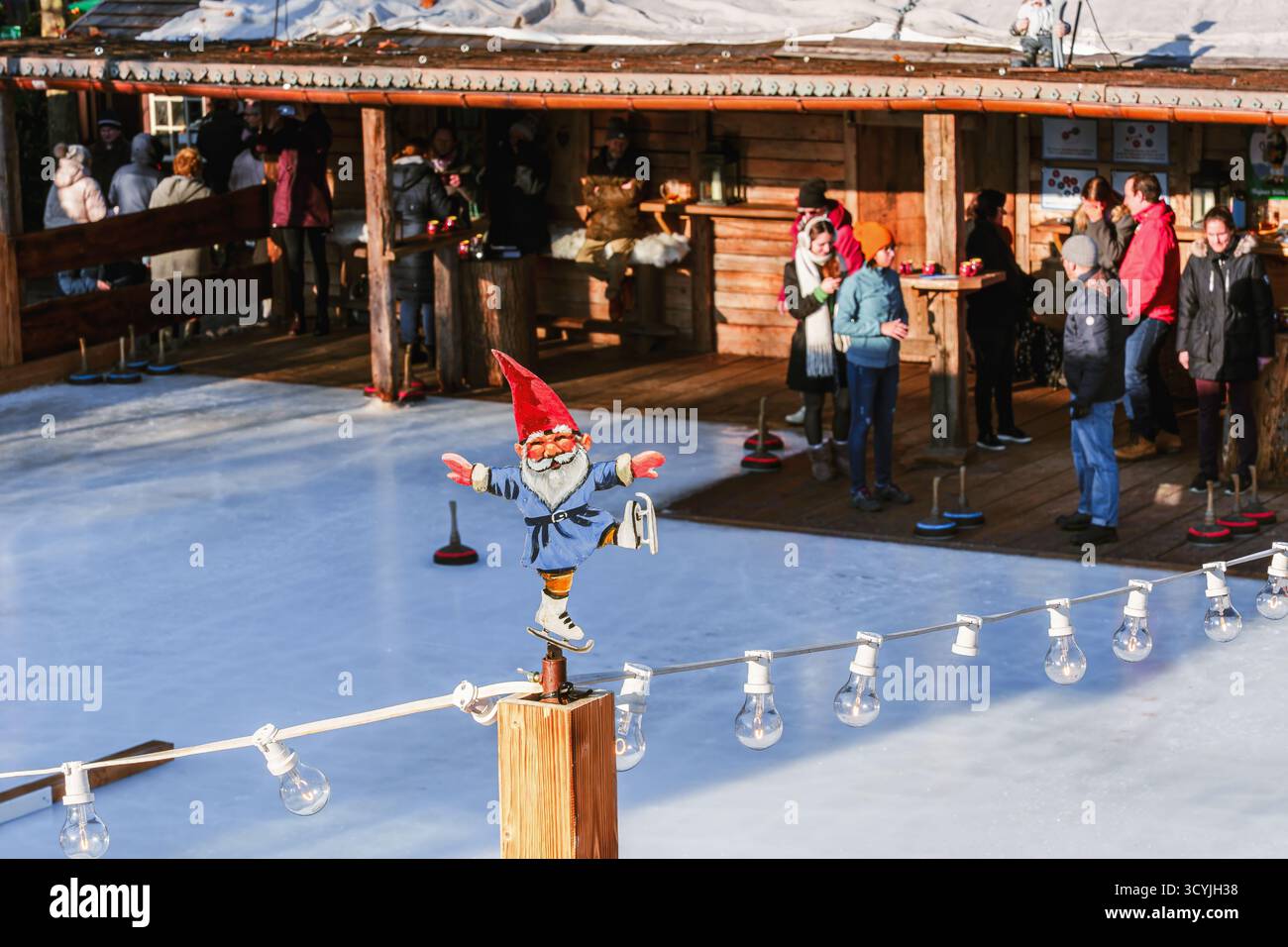 Köln, Deutschland - 12. Dezember 2022: Lustige Gnomenstatue in der Nähe einer Eisstockbahn, wo die Leute spielen und warme Getränke bei einem Holzwint genießen Stockfoto