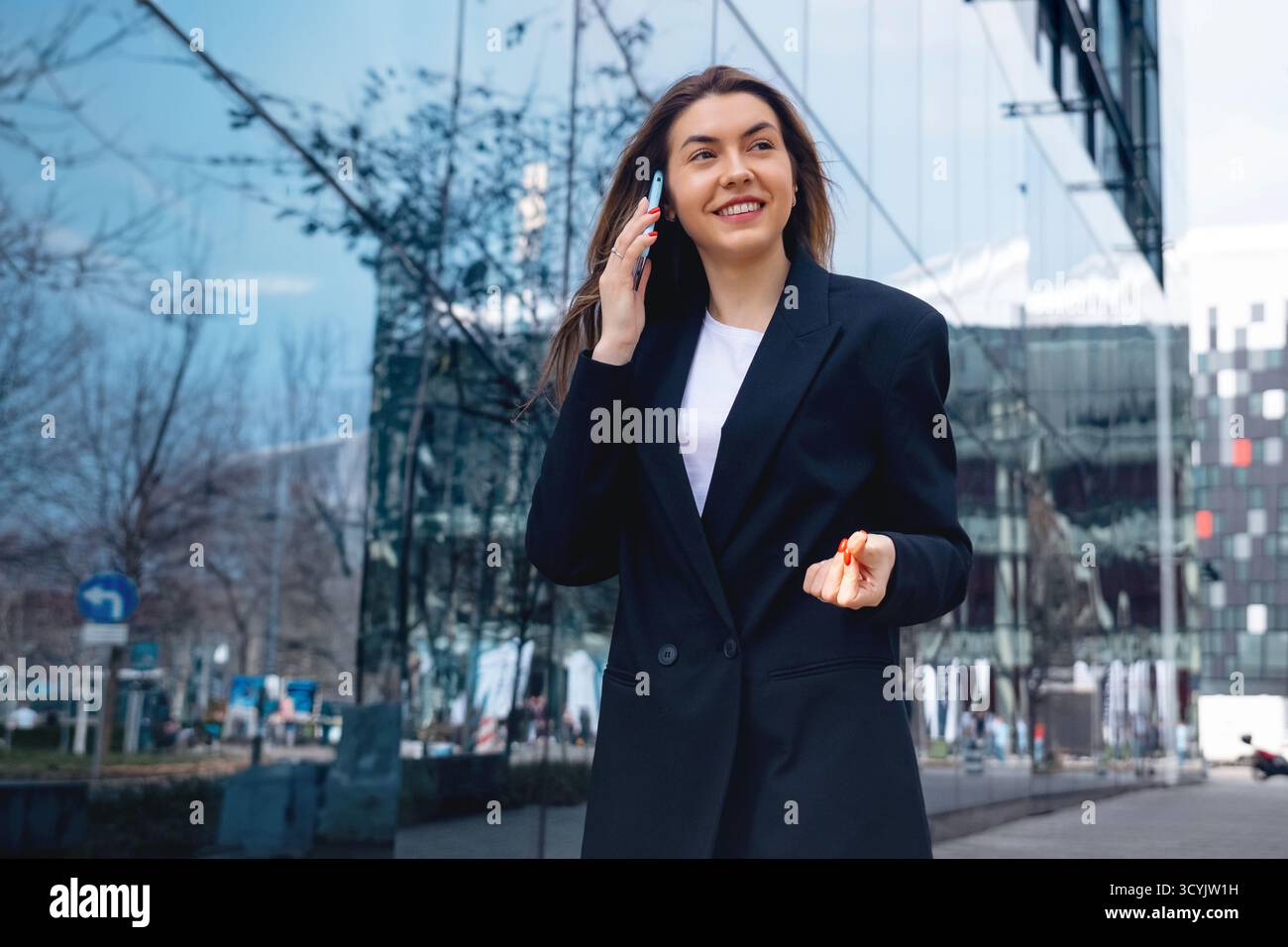 Eine selbstbewusste Geschäftsfrau läuft schnell, während sie am Telefon spricht. Sie trägt ein elegantes Outfit, umgeben von Bäumen und urbanen Lichtbögen Stockfoto