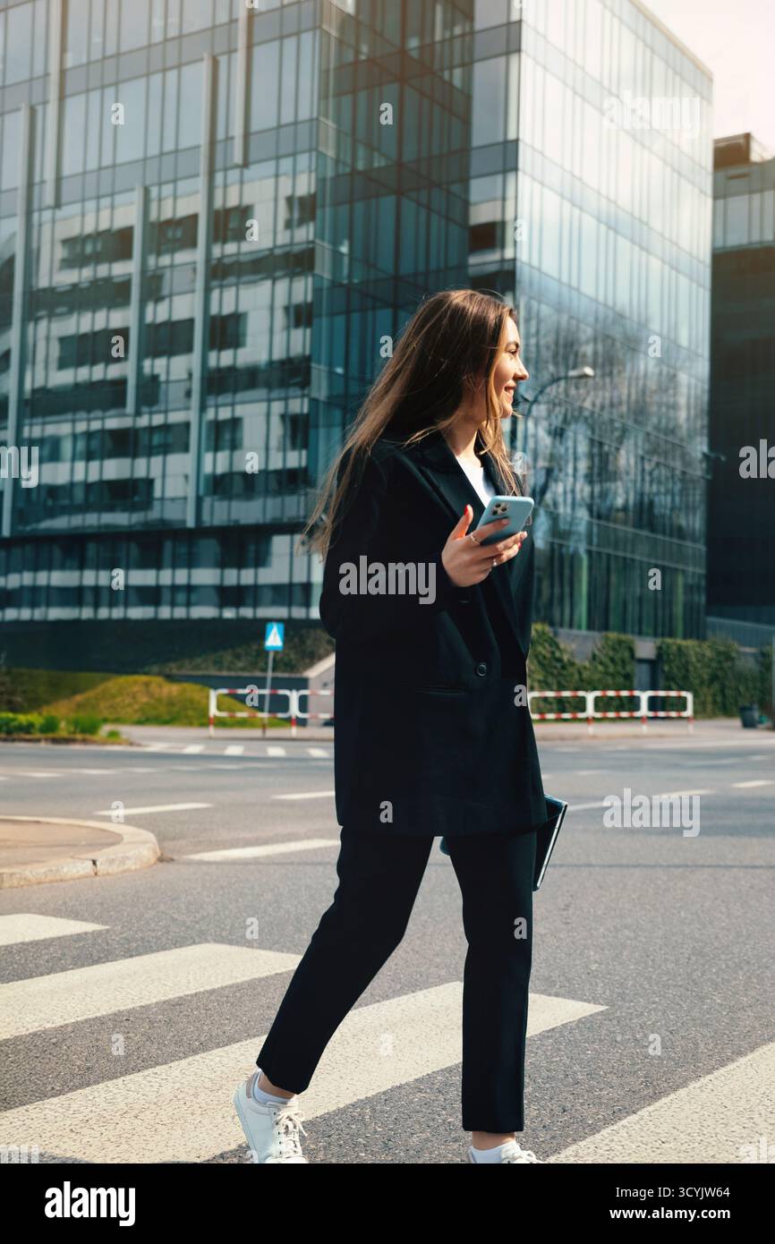 Eine junge Frau in stilvollem Outfit streift mit einem Smartphone in der Hand über einen Stadtübergang, umgeben von modernen Glasgebäuden auf einer hellen Straße Stockfoto
