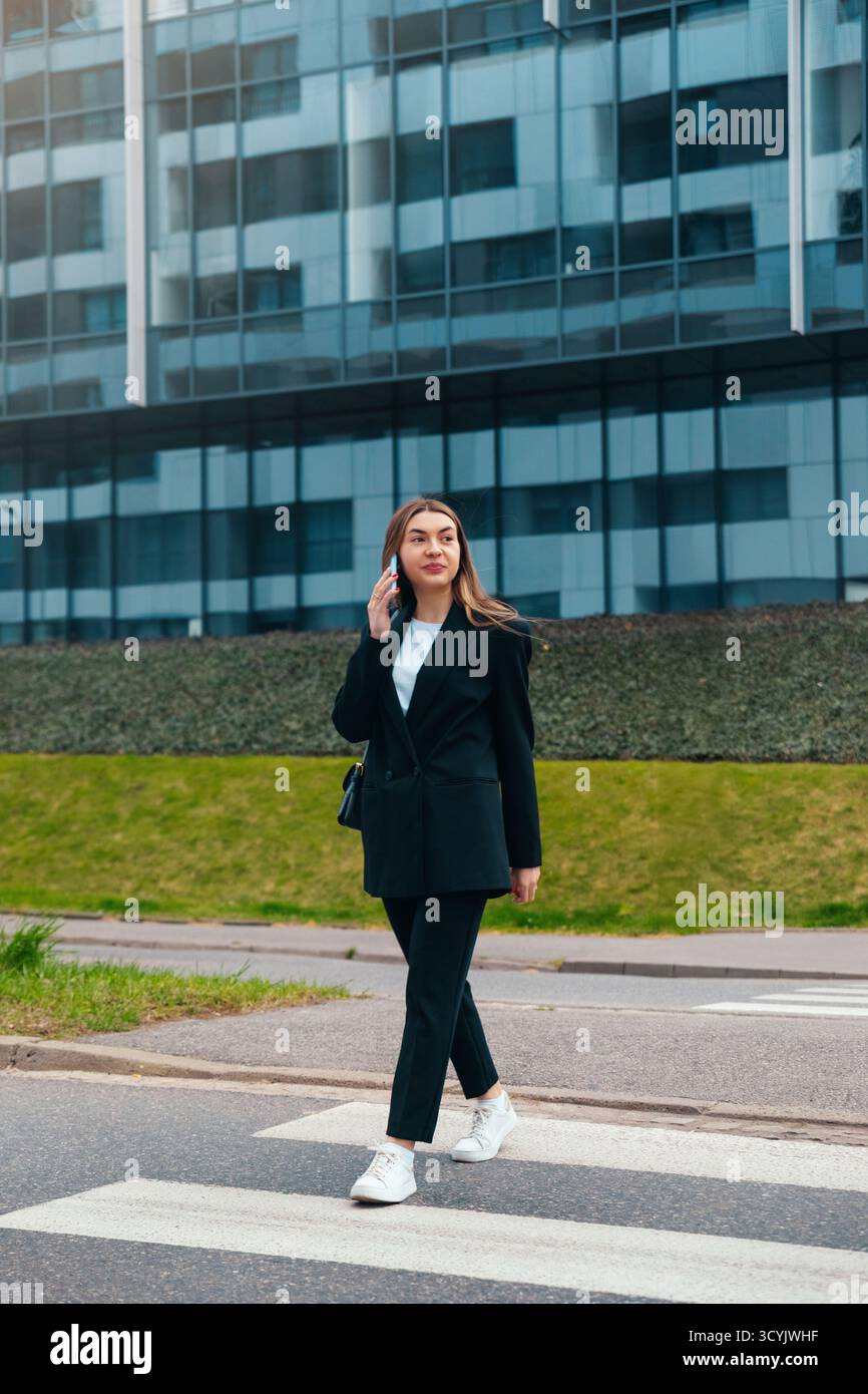 Eine Frau in einem schicken Outfit geht den Bürgersteig entlang, spricht selbstbewusst mit ihrem Telefon, vorbei an einem zeitgenössischen Glasgebäude i Stockfoto
