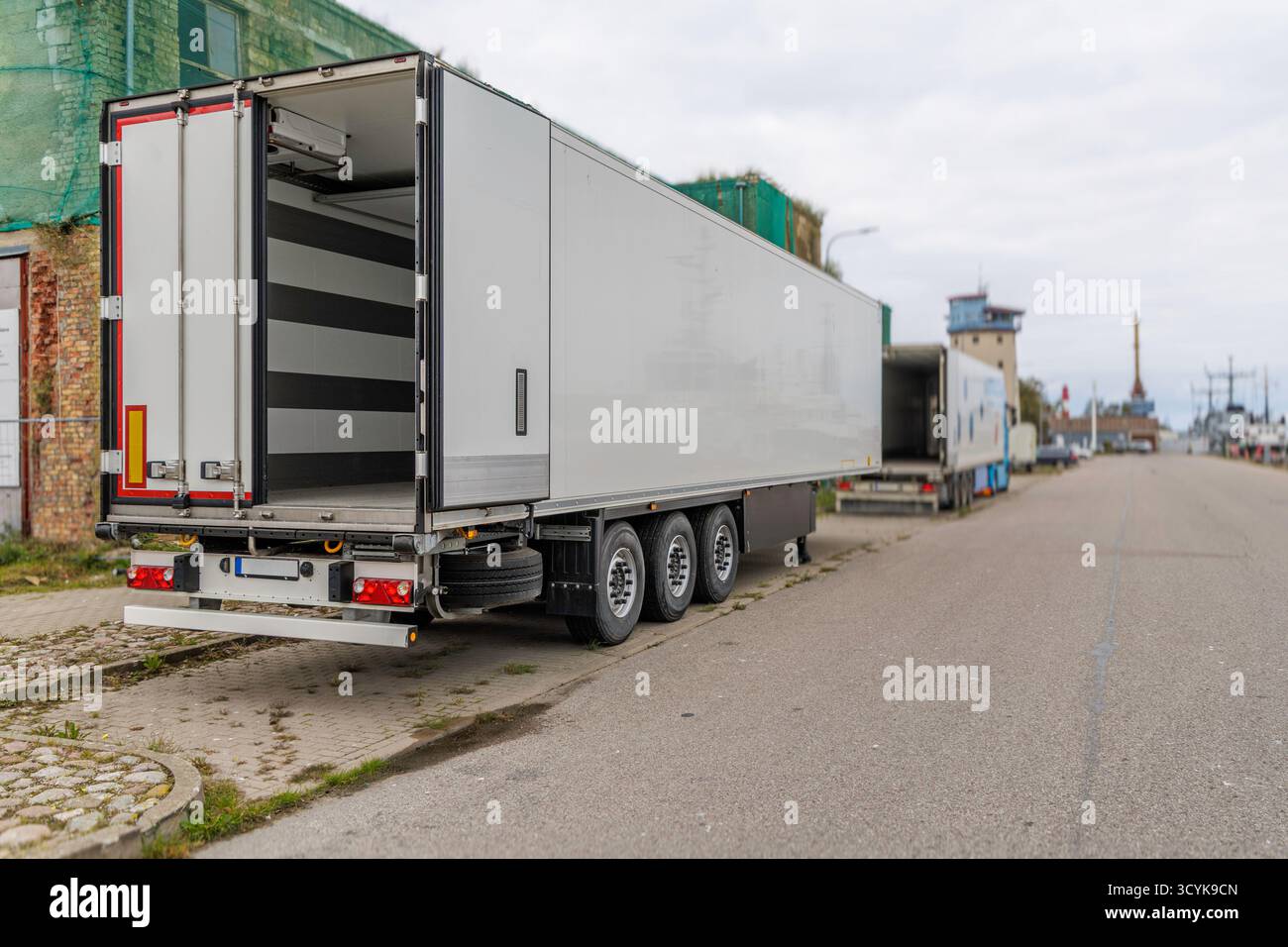 Die Heck- und Seitenansicht eines großen weißen Kühlsattelaufliegers mit offenen Türen, der auf der asphaltierten Straße eines Industriehafens auf einer bewölkten Fläche geparkt ist Stockfoto