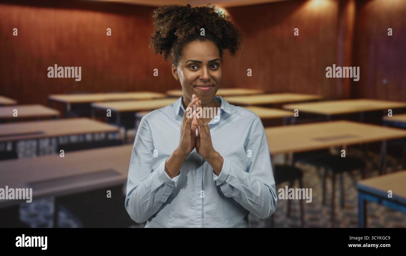 Frau mit gefesselten Händen und Fingern in einem Klassenzimmer zwischen leeren Schreibtischen, mit einem leichten Hemd und lächelnd; Unfug. Stockfoto