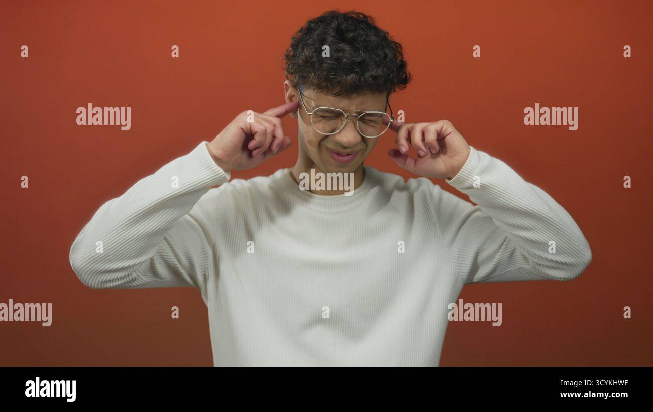 Mann mit Brille, lockigen Haaren und weißem Hemd schaut mit gebeugter Haltung gegen die orangene Studiowand; Nachsicht. Stockfoto
