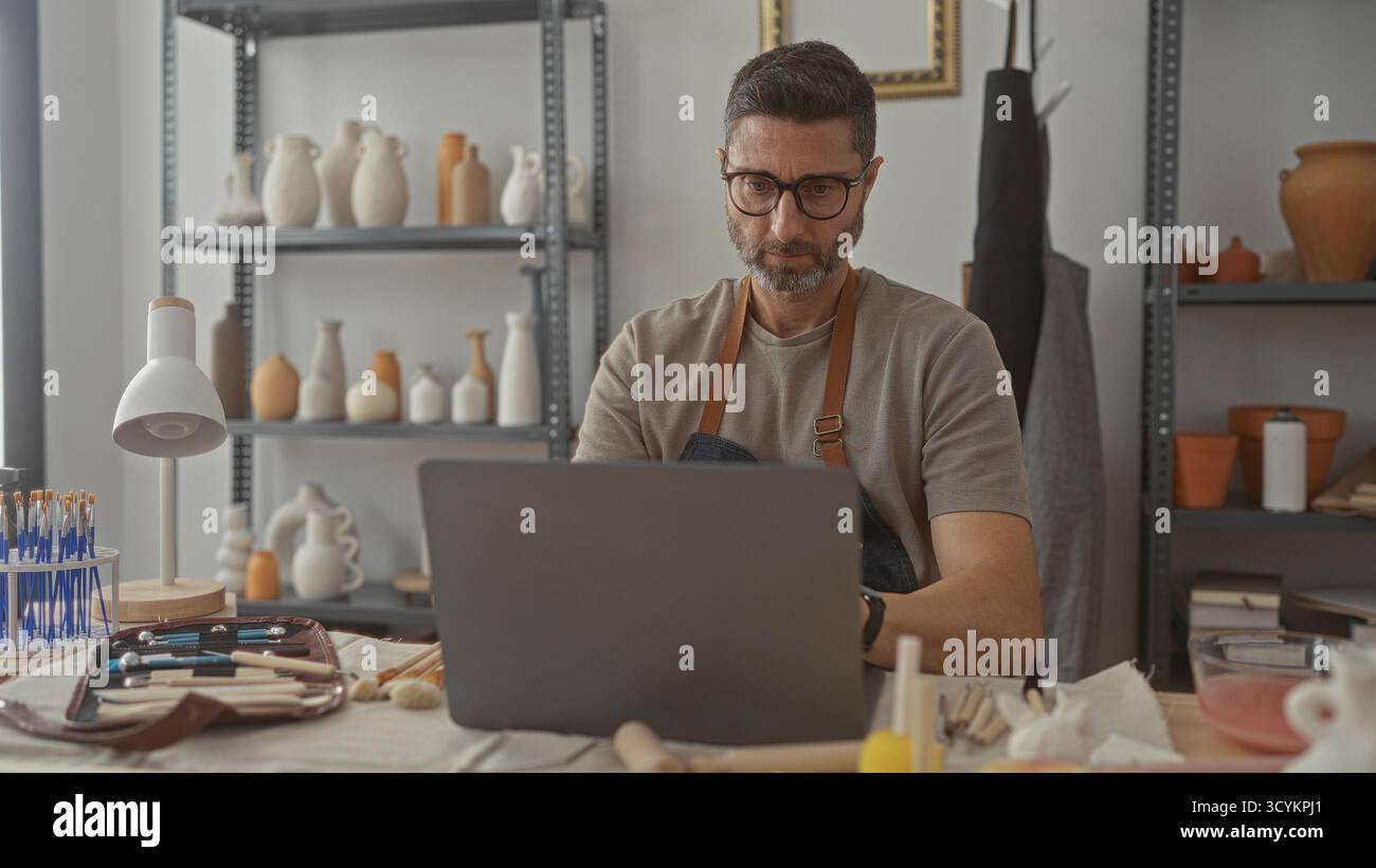 Mann Töpfer mit Hand am Kinn mit Laptop im Studio zwischen Töpferregalen, Bürsten und Tonwerkzeugen; durchdachte Handwerkspraxis. Stockfoto
