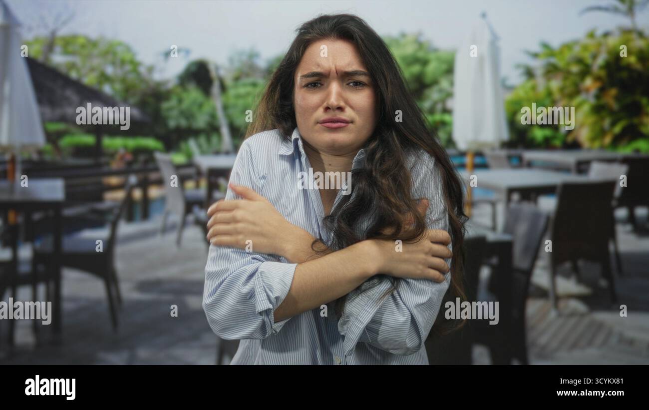 Die junge hispanische Frau, die ihre Arme umarmt, zittert in einem Gebäude auf einer Restaurantterrasse mit geschlossenen Augen und angespannter Haltung; Unwohlsein. Stockfoto