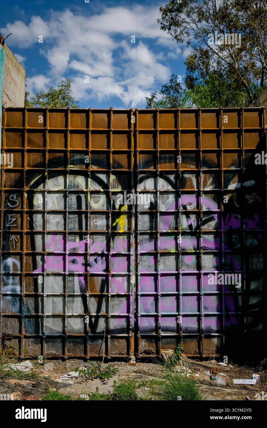 Städtische Graffiti-Kunst auf einer Metallgitterwand, umgeben von Grün unter einem klaren blauen Himmel mit verstreuten Wolken Stockfoto