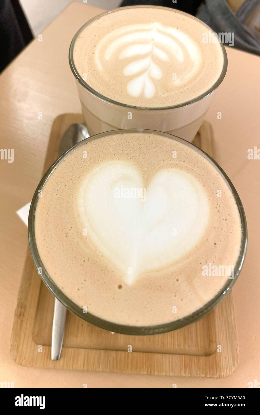 Zwei wunderschön gefertigte Kaffeegetränke mit komplizierten Latte Art Designs, eines mit Herz und das andere mit Blattmuster, präsentiert auf einem Holztablett Stockfoto