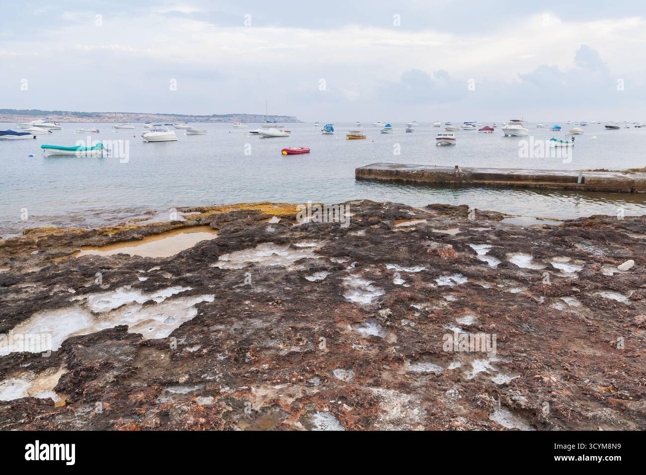 Eine felsige Küste führt zu einem ruhigen Hafen voller kleiner Boote. Sanftes Licht, bedeckter Himmel und stilles Wasser schaffen eine friedliche Küstenatmosphäre o Stockfoto