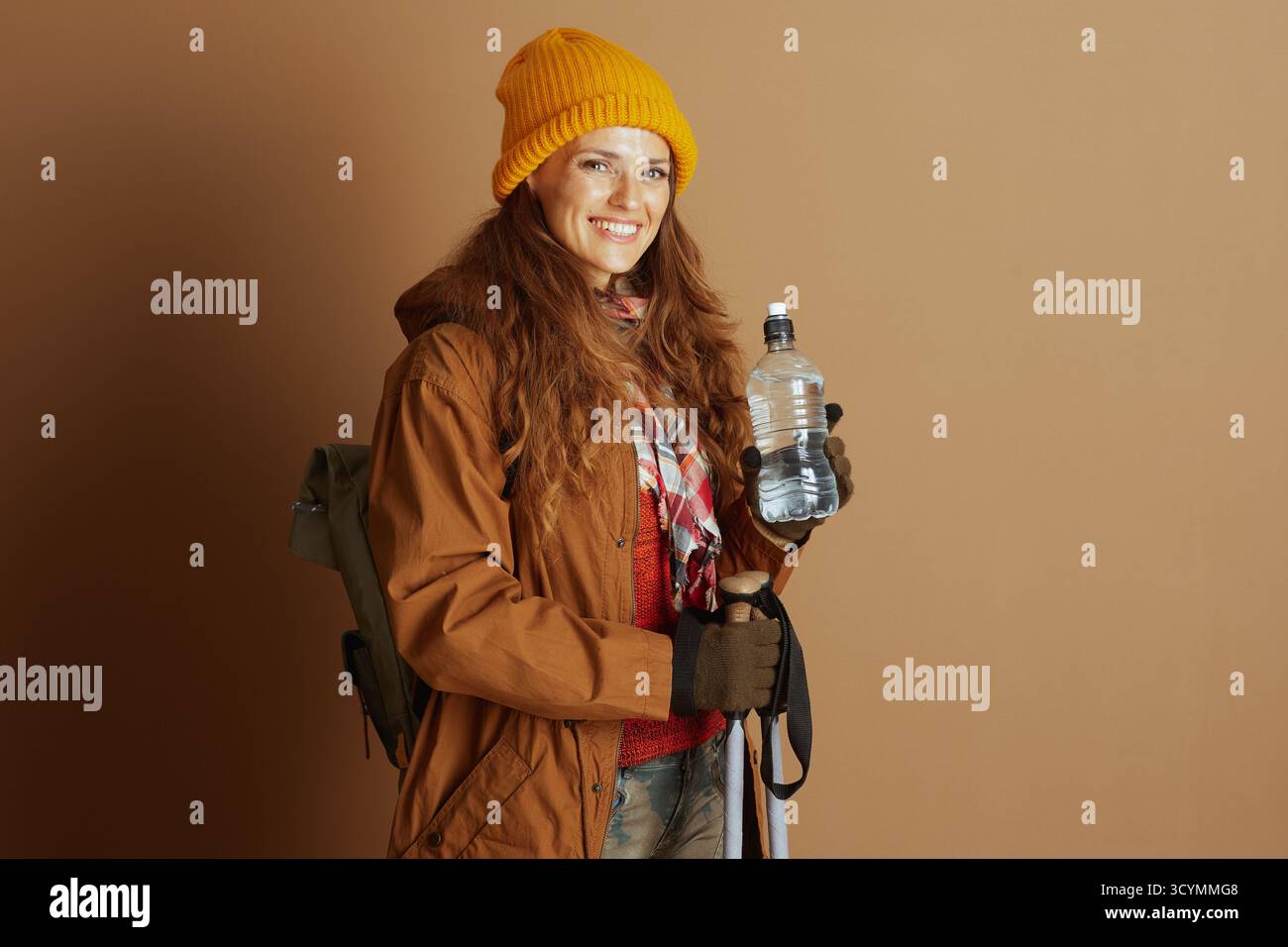 Eine lächelnde junge Erwachsene Frau in den späten 20er oder frühen 30er Jahren, die gelbe Beanie, braune Kapuzenjacke und blaue Jeans trägt, steht mit Rucksack, Wasserflasche und Stockfoto