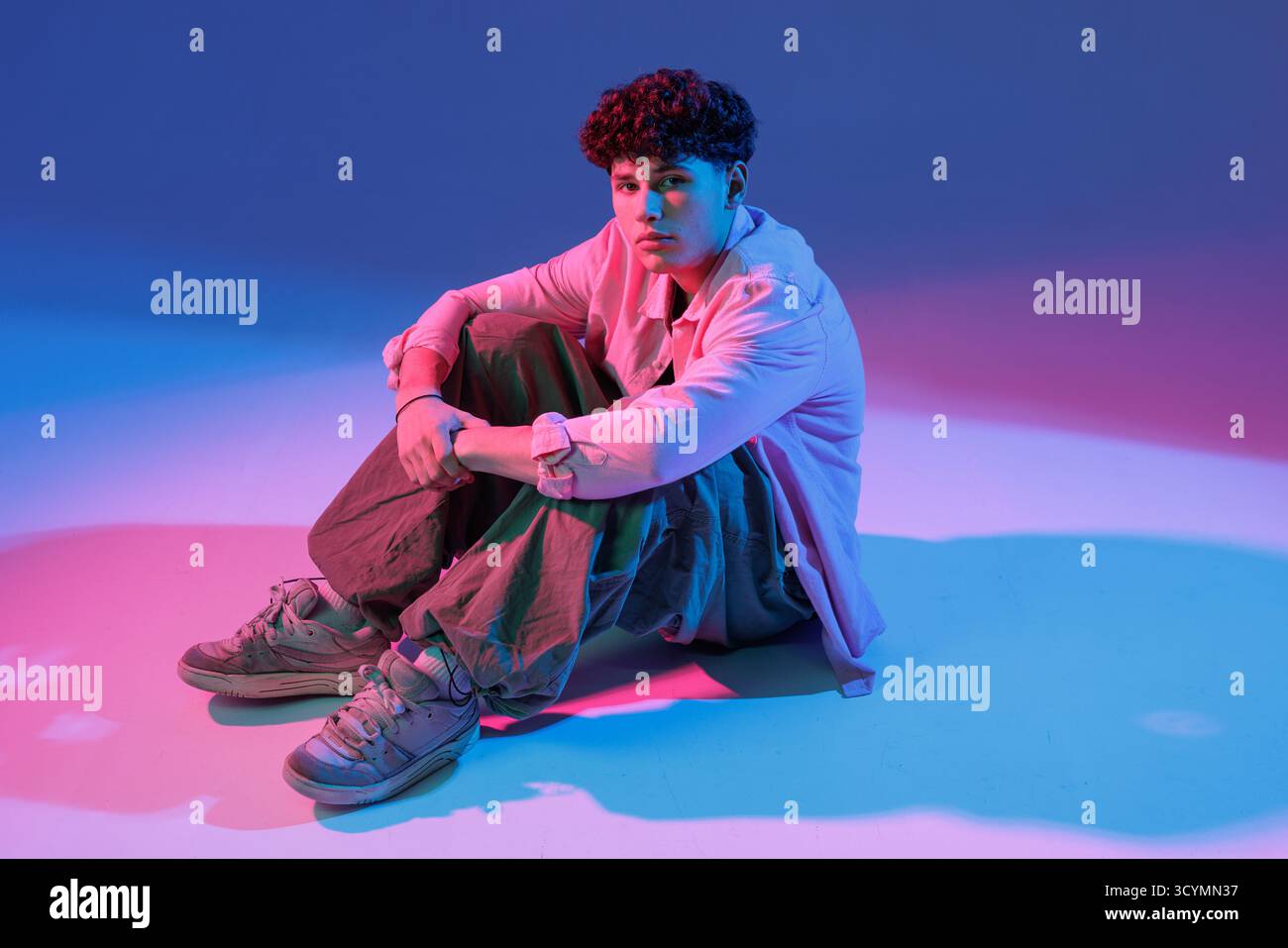 Junge Tänzerin in Baggy Streetwear sitzt unter pinkblauen Neongelen, stilvollem Hip-Hop-Porträt, bereit für Musik, Mode und Event-Promotionen. Stockfoto