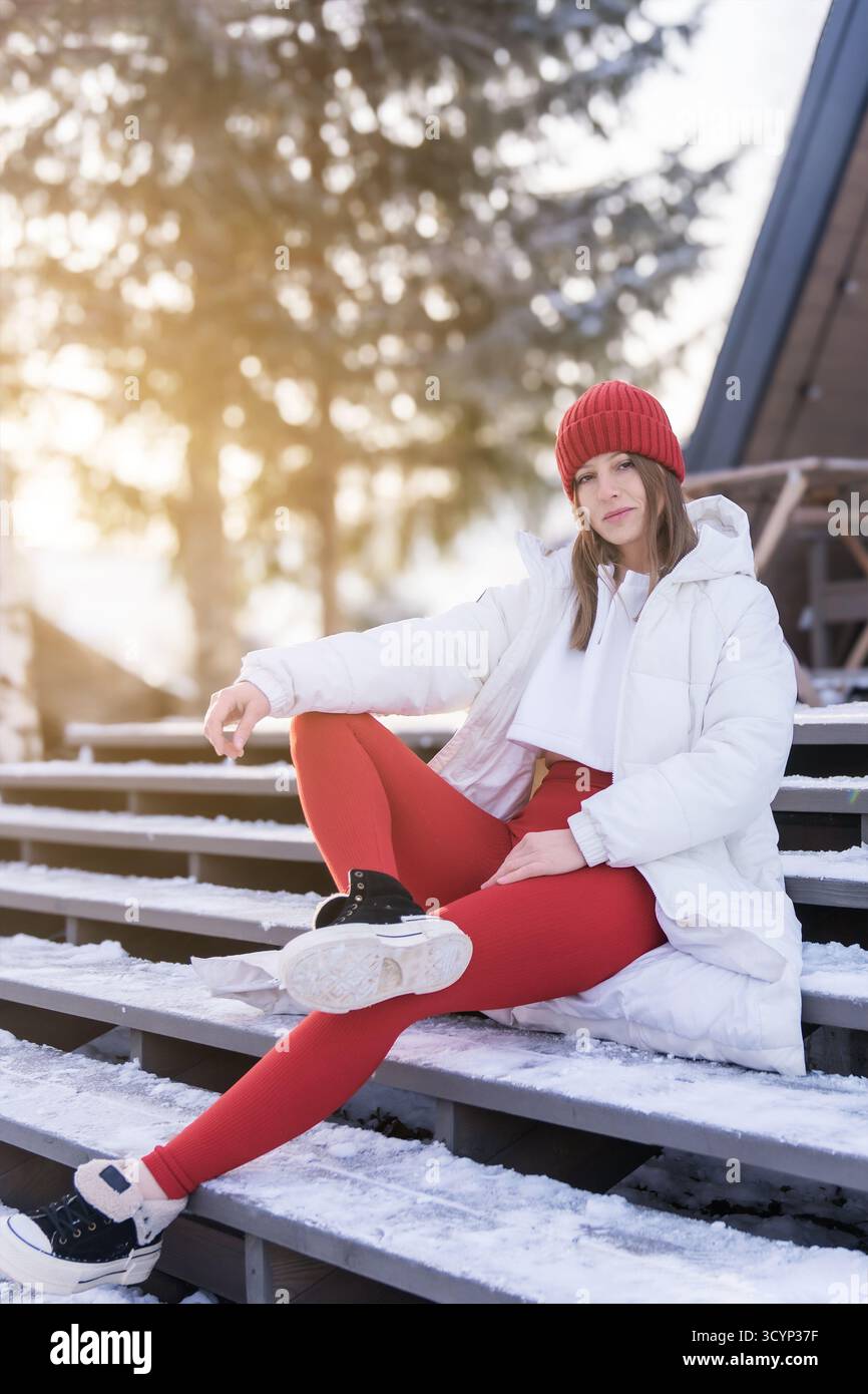 Junge Frau in Wintermode, die sich bei Tageslicht auf verschneiten Stufen in sonniger Umgebung im Freien entspannt Stockfoto