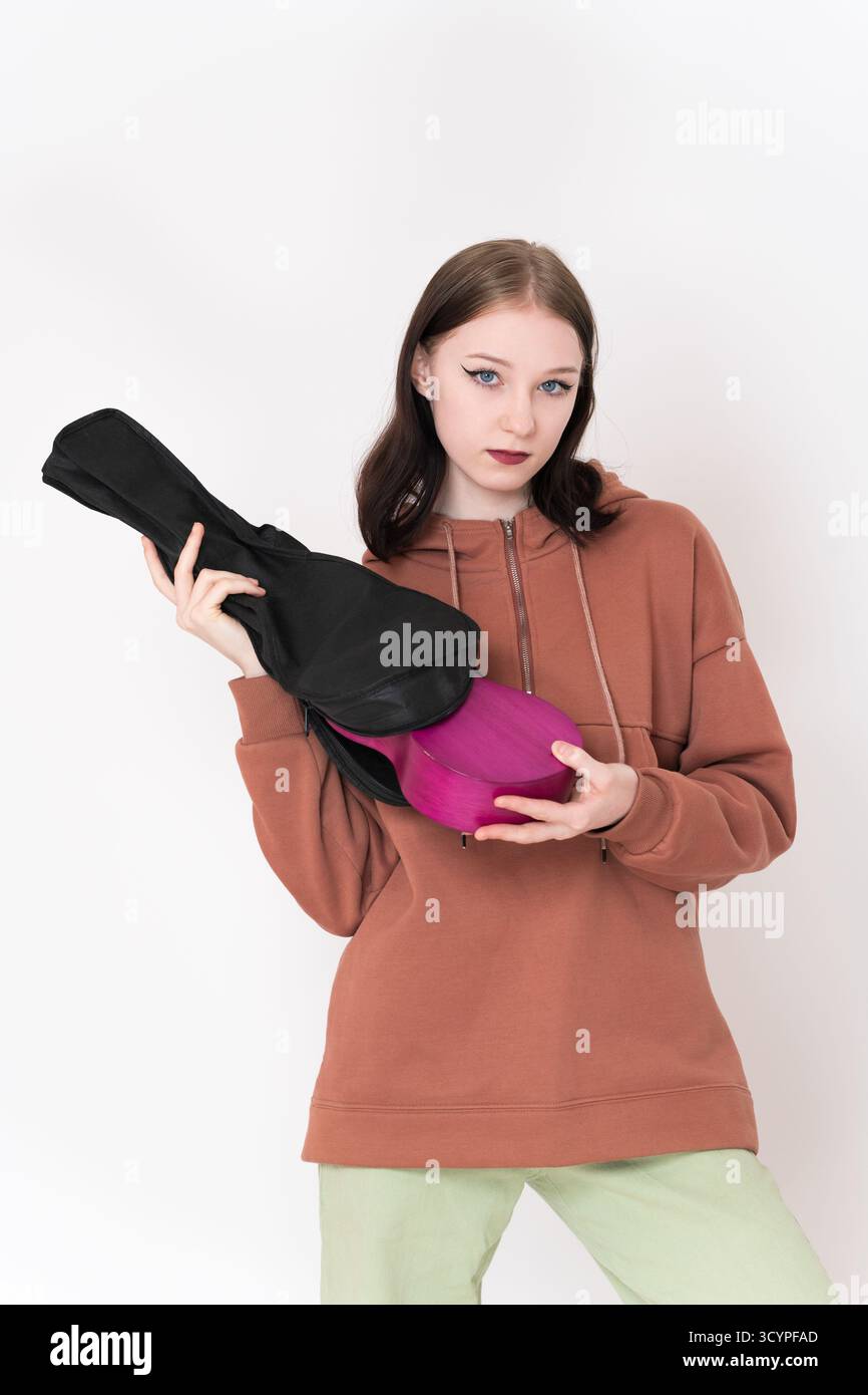 Die Ukulele-Musikerin nimmt die Ukulele aus der Tasche und schaut in die Kamera. Authentische Frau in braunem Hoodie und hellgrüner Jeans Stockfoto