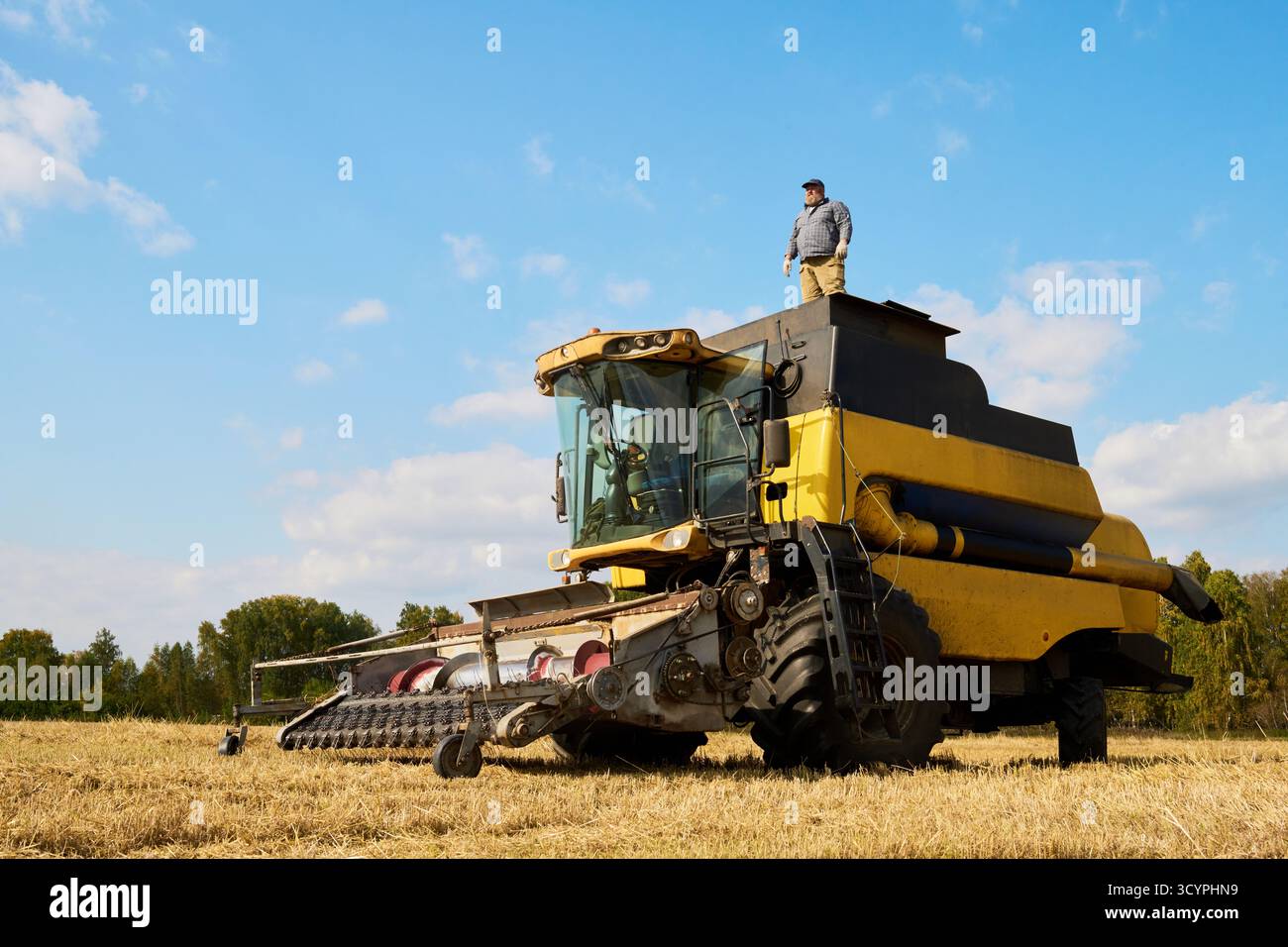 Kaukasischer Mann mittleren Alters, der während der Erntesaison auf einem großen landwirtschaftlichen Mähdrescher auf offenem Feld steht, Ackerland beobachtet und den Maschinenbetrieb überwacht Stockfoto