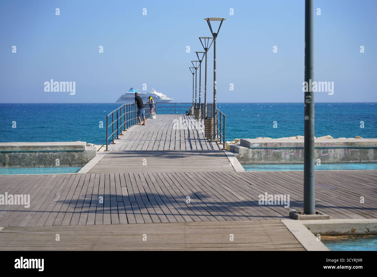 Paare genießen einen gemütlichen Spaziergang auf einem sonnigen Pier mit Blick auf das ruhige blaue Wasser im Mittelmeerraum Stockfoto