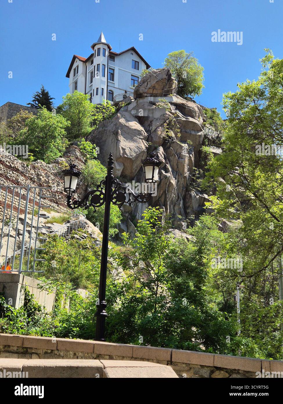 Das Foto zeigt einen hohen felsigen Hügel, auf dem ein helles historisches Gebäude mit einem dunklen Dach steht. Der Hügel ist mit großen grauen Felsen und bedeckt - Smartphone-aufgenommenes Stockfoto