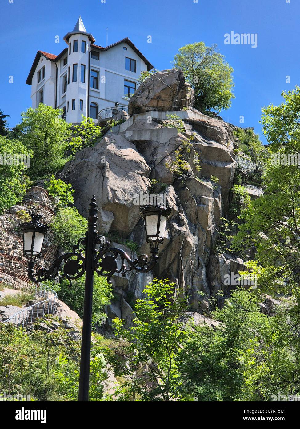 Das Foto zeigt einen hohen felsigen Hügel, auf dem ein helles historisches Gebäude mit einem dunklen Dach steht. Der Hügel ist mit großen grauen Felsen und bedeckt - Smartphone-aufgenommenes Stockfoto