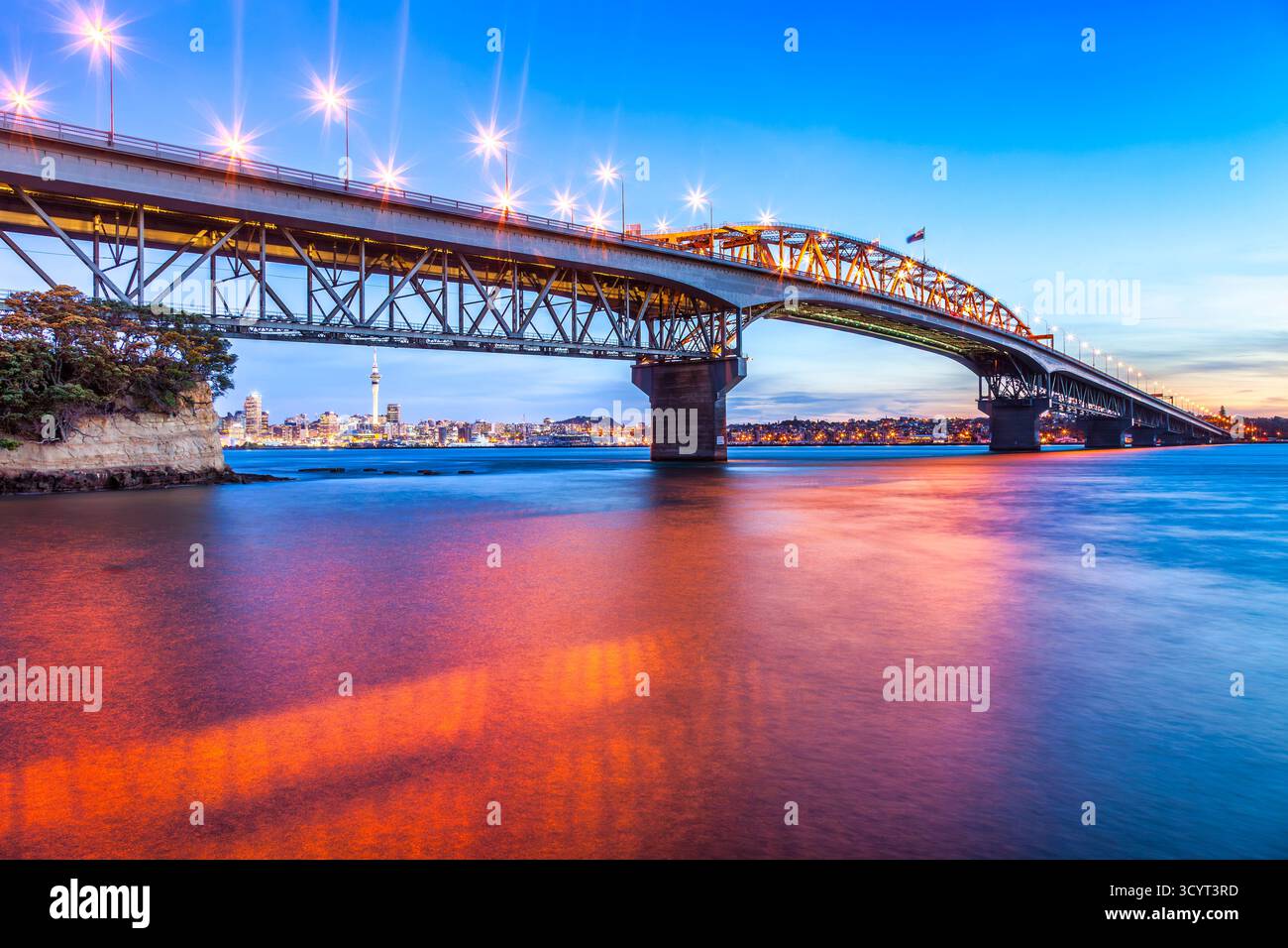 Auckland Harbour Bridge bei Dämmerung, mit leuchtenden Lichtern und roter Reflexion im Hafen. Stockfoto