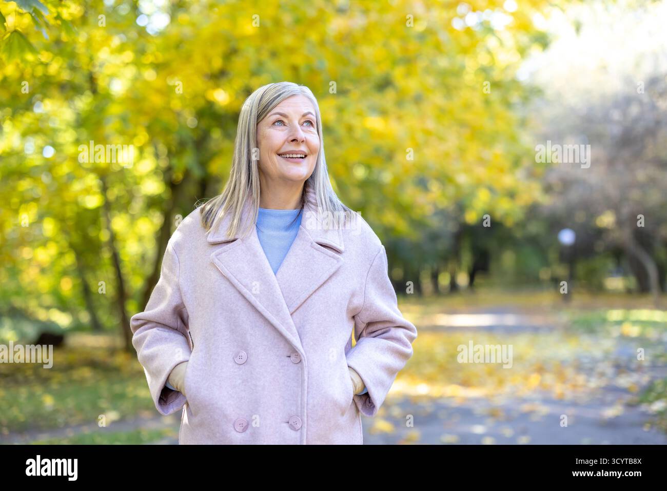 Eine ältere Frau in einem stilvollen Mantel lächelt und blickt auf und genießt einen gemütlichen Spaziergang voller goldener Blätter in einem wunderschönen, sonnendurchfluteten Herbstpark Stockfoto