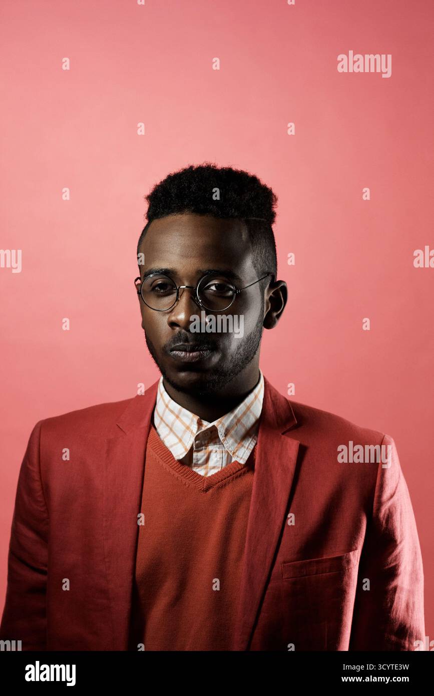 Porträt eines jungen, modischen afrikanischen Mannes, der eine Brille trägt und in die Kamera blickt, während er in schwacher Beleuchtung vor rotem Hintergrund steht Stockfoto