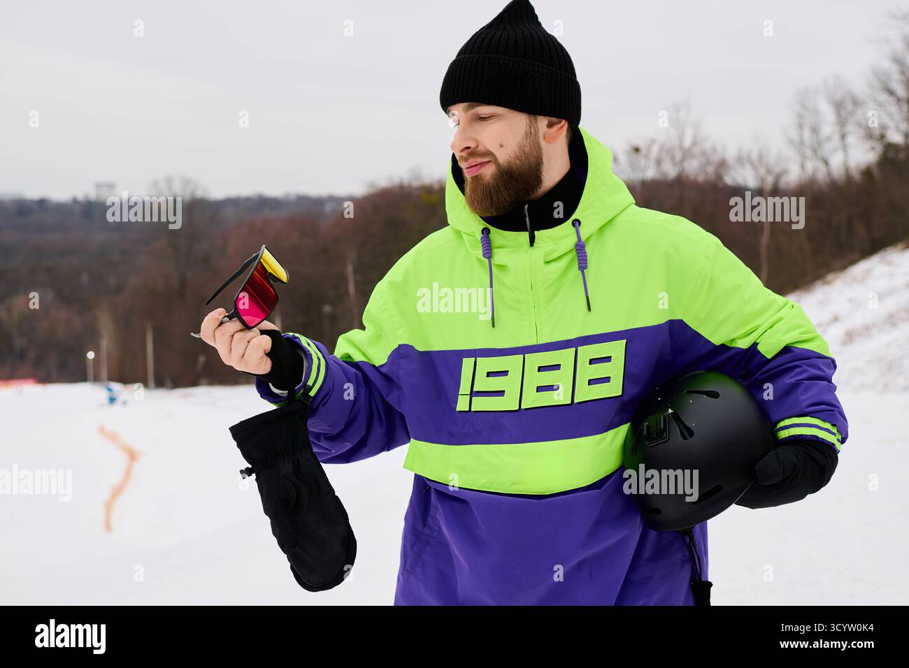 Ein bärtiger Mann hält beim Snowboarden auf einer verschneiten Piste inmitten von Winterlandschaft eine bunte Brille. Stockfoto
