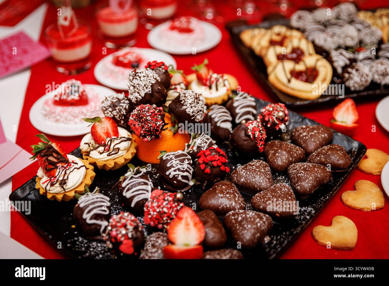 Verschiedene Erdbeeren mit Schokoladenüberzug, Herzkekse und Mini-Torten auf festlichem rotem Hintergrund. Ideal für Valentinstag, romantische Veranstaltung Stockfoto