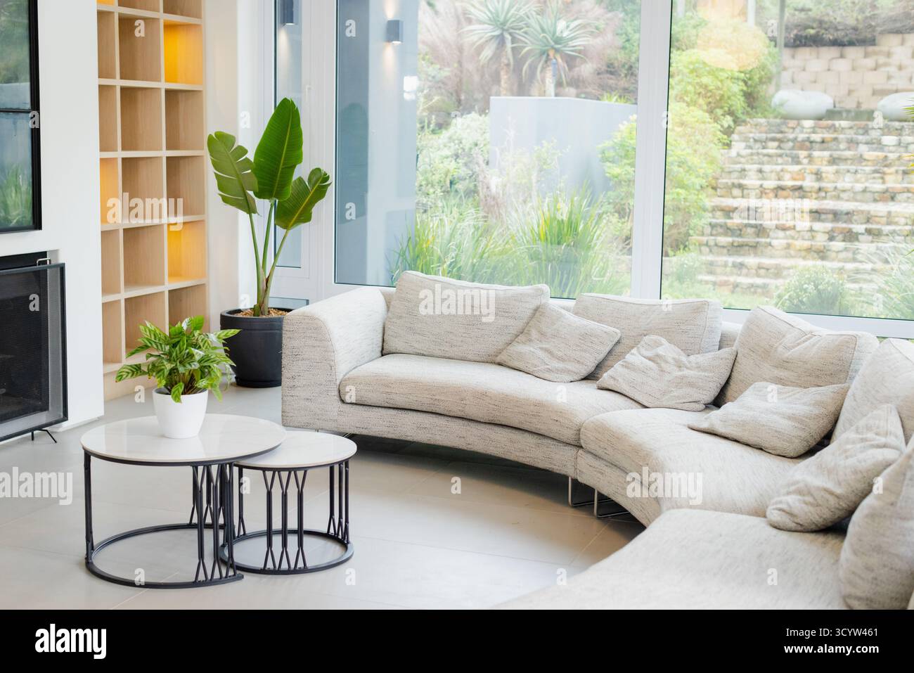 Das geschwungene hellgraue Sofabett ist an hohen Fenstern mit Couchtischen und Pflanzen befestigt Stockfoto