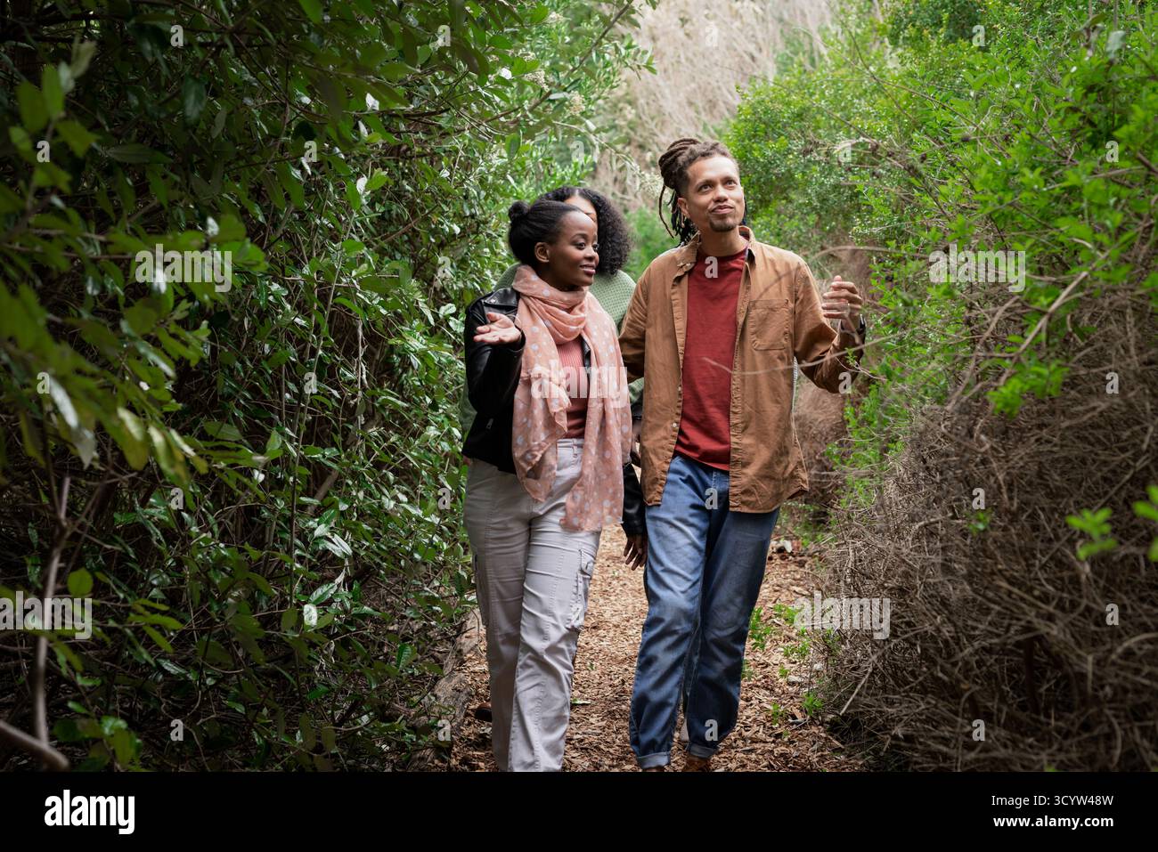 Abwechslungsreiche Paare, die auf einem Feldweg im Naturschutzgebiet wandern, umgeben von grünen Sträuchern und trockenen Gräsern Stockfoto