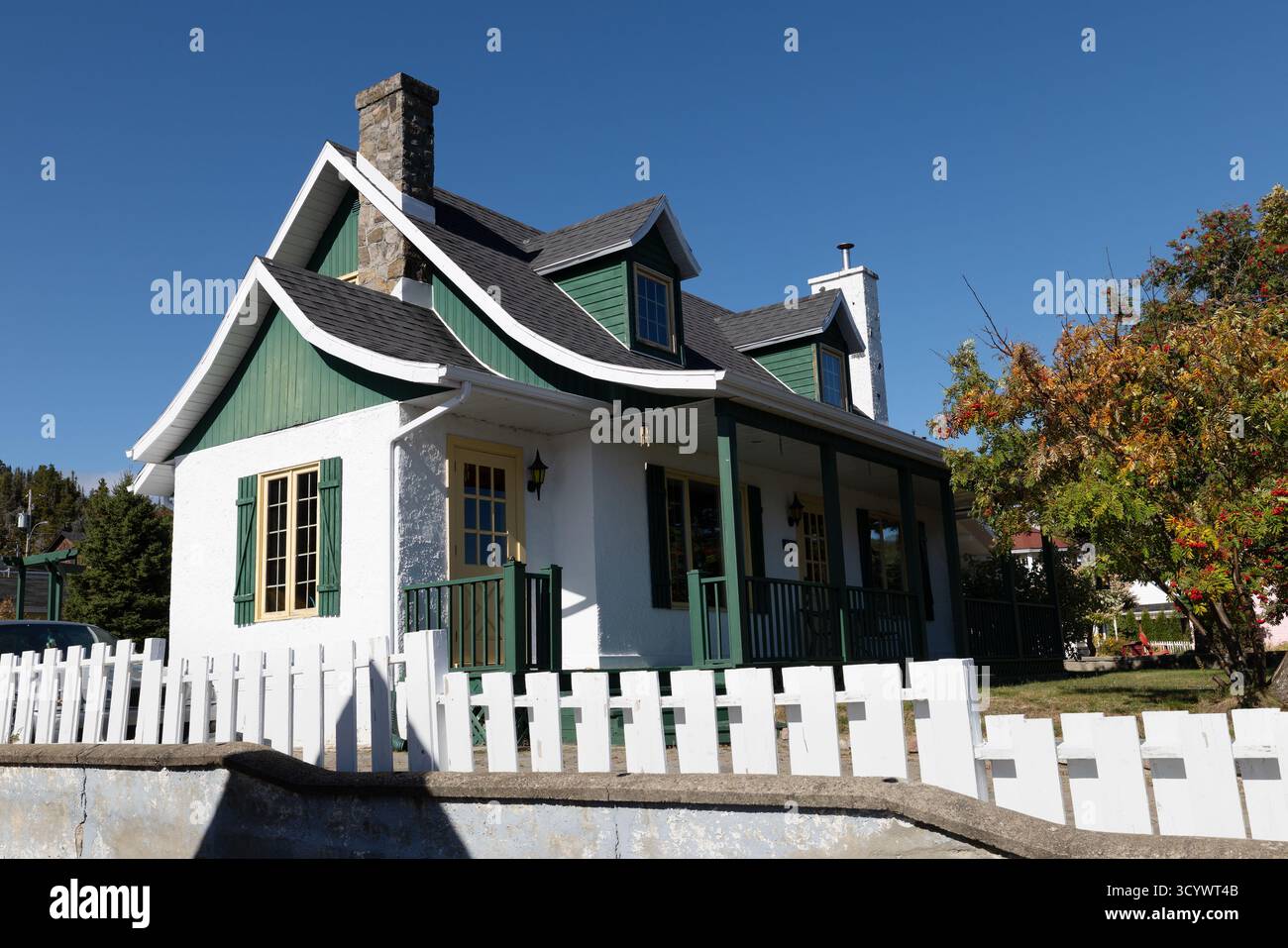 Charmantes traditionelles Haus mit grüner Zierleiste und weißem Pflückzaun unter einem klaren blauen Himmel in Tadoussac, Quebec, Kanada. Stockfoto