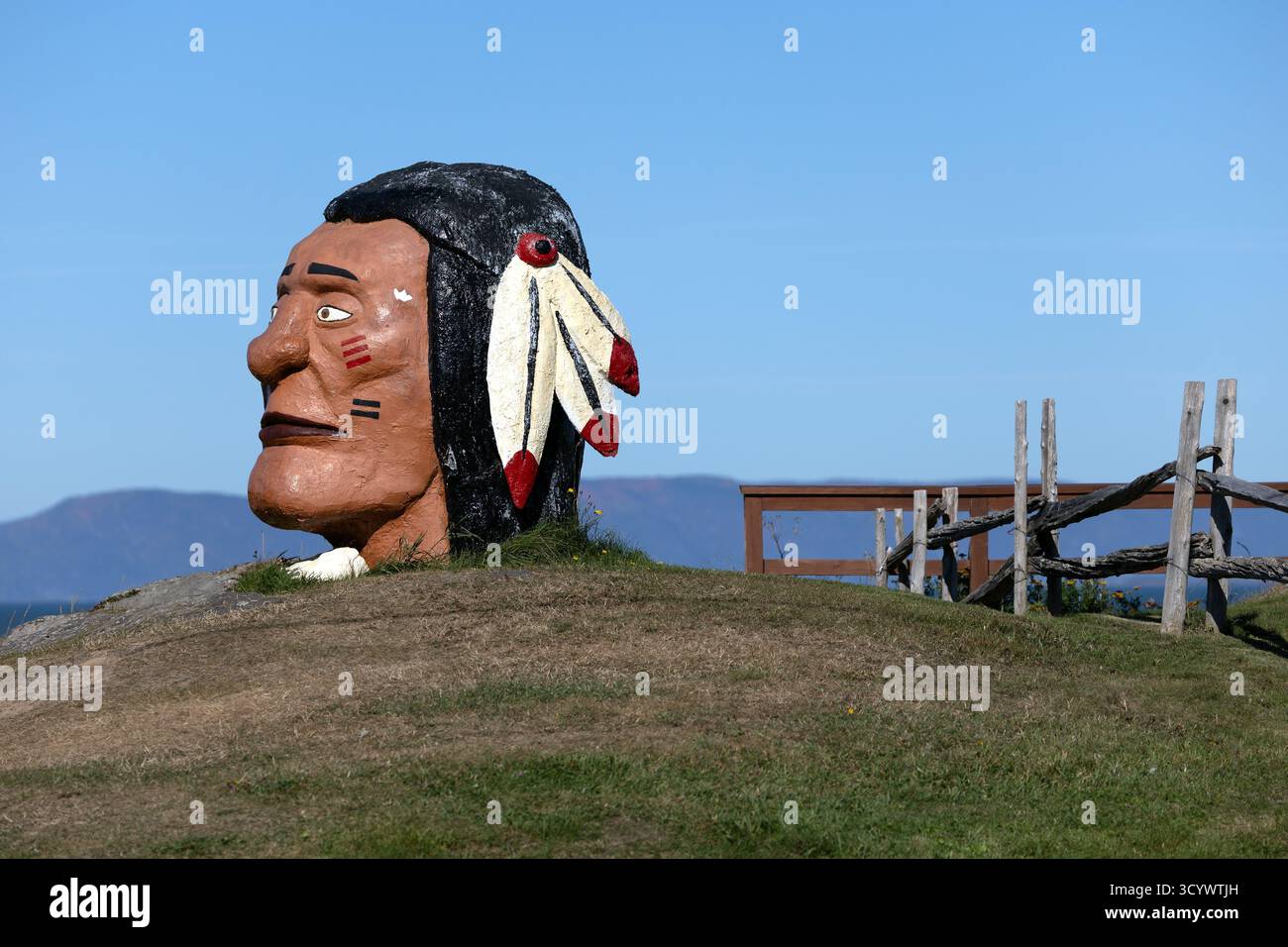 Eine große gemalte Skulptur eines menschlichen Kopfes mit stilisierten Federn steht auf einem grasbewachsenen Hügel unter einem klaren blauen Himmel, Rivière-du-Loup, Quebec. Stockfoto
