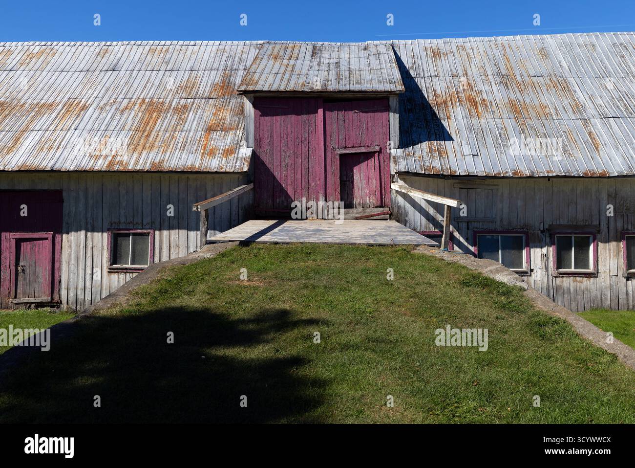 Eine alte Holzscheune mit verblassten roten Türen und einem rostigen Blechdach steht unter einem klaren blauen Himmel in Quebec. Stockfoto