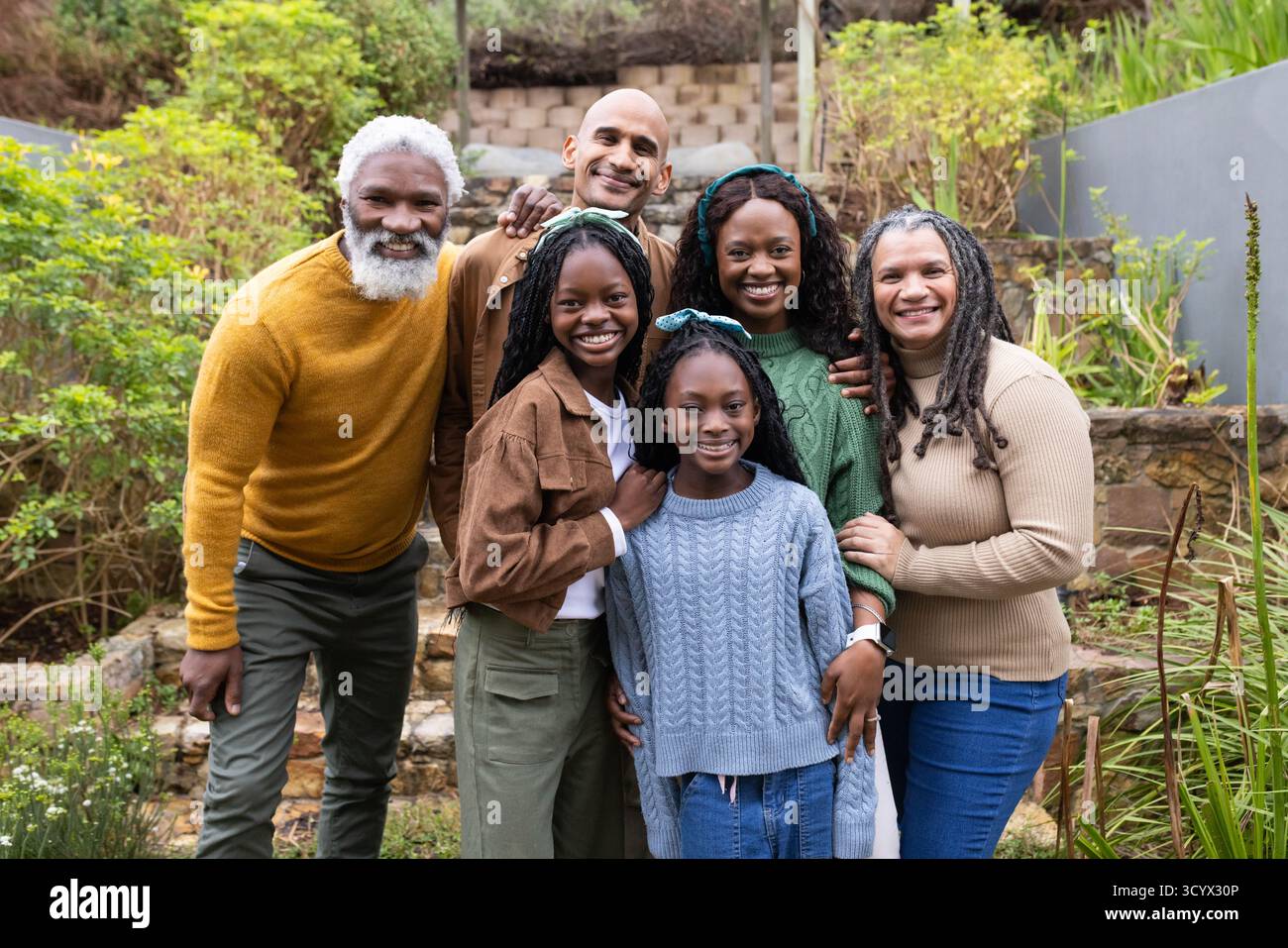 Vielfältige Familie, die auf Stufen im Garten unter der Pergola mit gestrickten Haarbändern steht und den Betrachter anlächelt Stockfoto