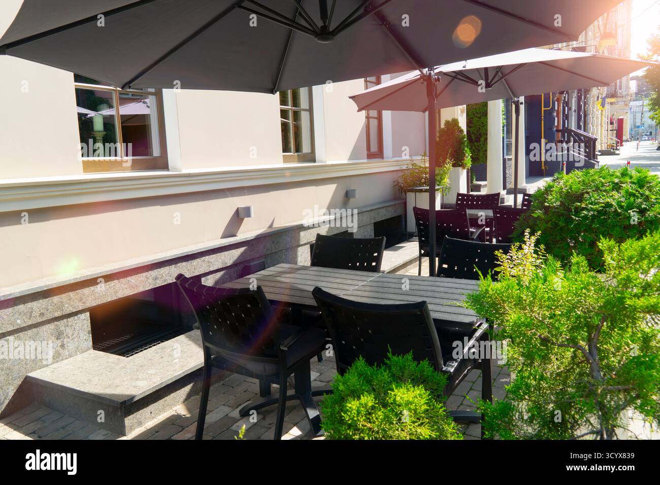 Sommerblick auf ein leeres Straßencafé oder eine Restaurantterrasse mit dunklen Tischen und Stühlen, die von großen schwarzen Sonnenschirmen im hellen Sonnenlicht beschattet werden Stockfoto