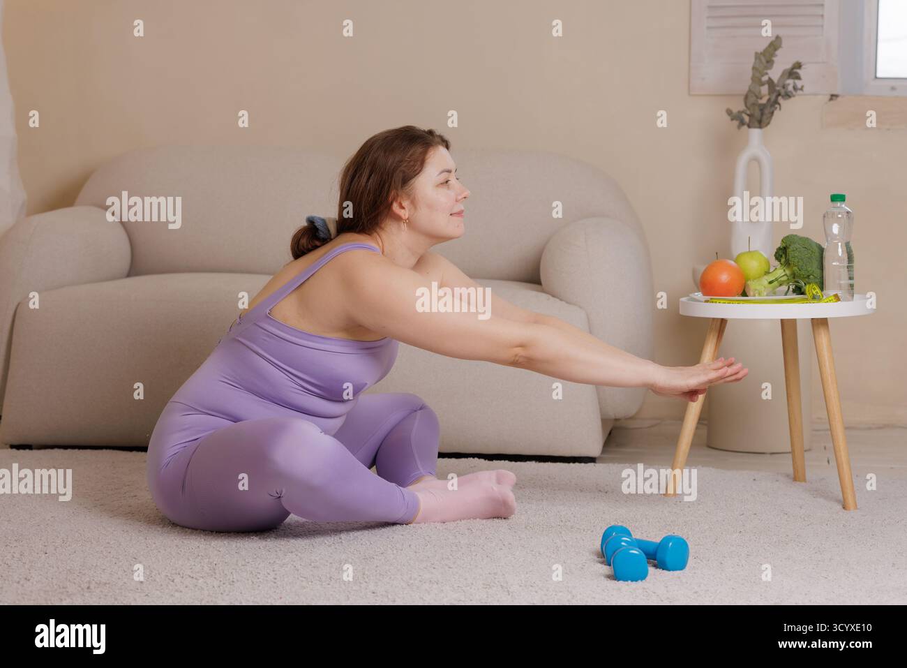 Positive Home Workout Szene einer Plus Size Frau dehnt sich mit Kurzhanteln Obst Gemüse und Wasser hebt Gewichtskontrolle und gesunde Routine hervor Stockfoto