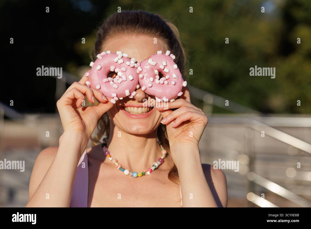Fröhliche Frau bedeckt die Augen mit rosafarbenen Donuts mit Marshmallows verspieltes Dessertporträt im Freien bei warmem Sonnenlicht Stockfoto