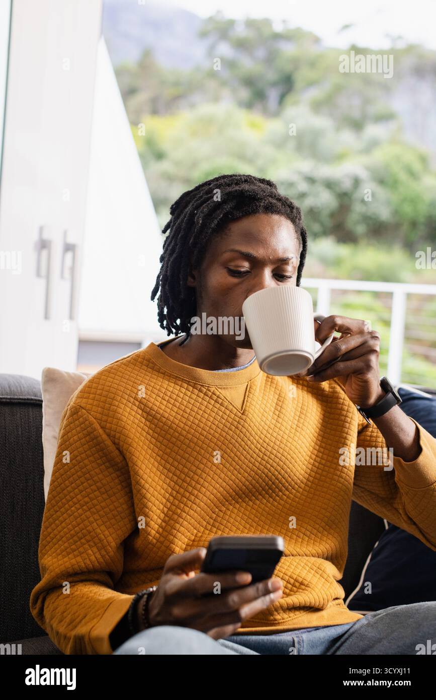 Afroamerikaner sitzt auf dem Sofa, trägt Sweatshirt und Smartwatch, hält Becher und Smartphone Stockfoto