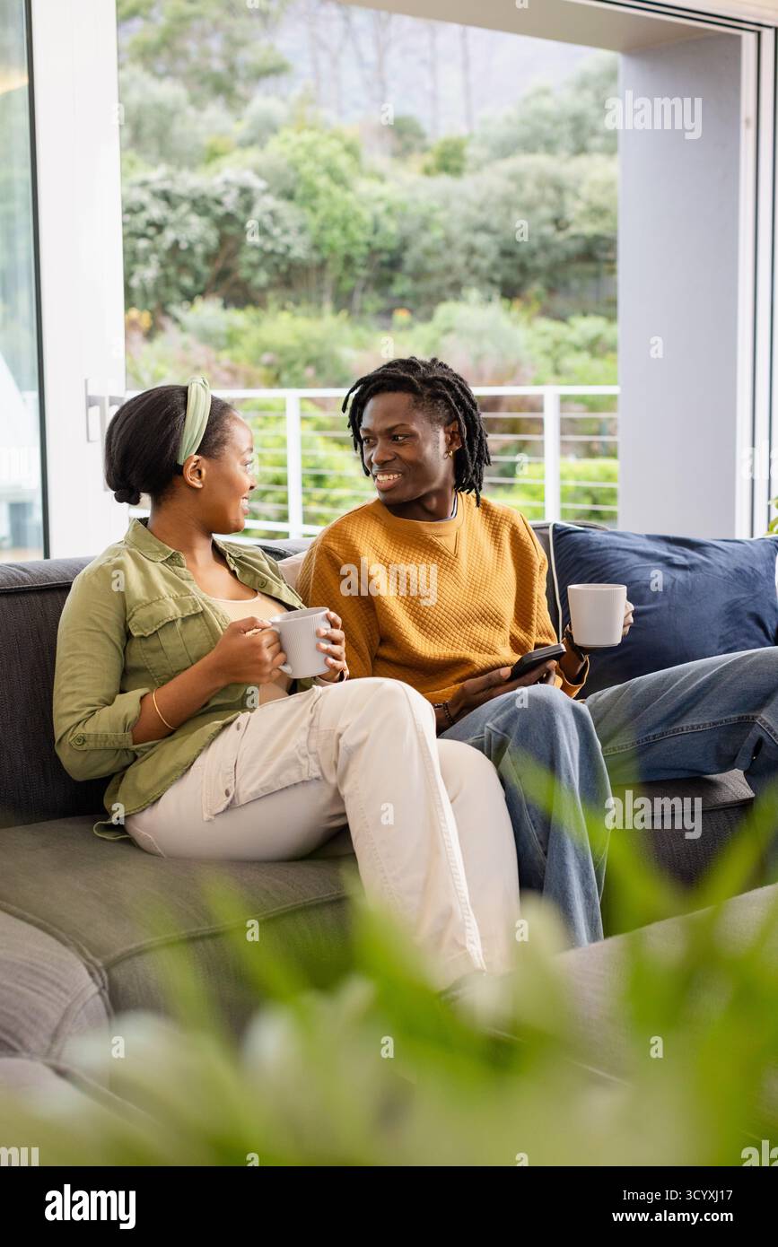 Afroamerikanisches Paar unterhält sich auf einem dunkelgrauen Sofa im Wohnzimmer mit Keramikbecher und Tablet Stockfoto