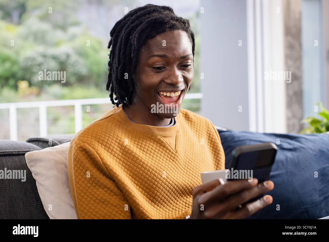 Afroamerikaner, der Smartphone und Becher hält, während er auf dem Sofa am Fenster mit Kissen lächelt Stockfoto