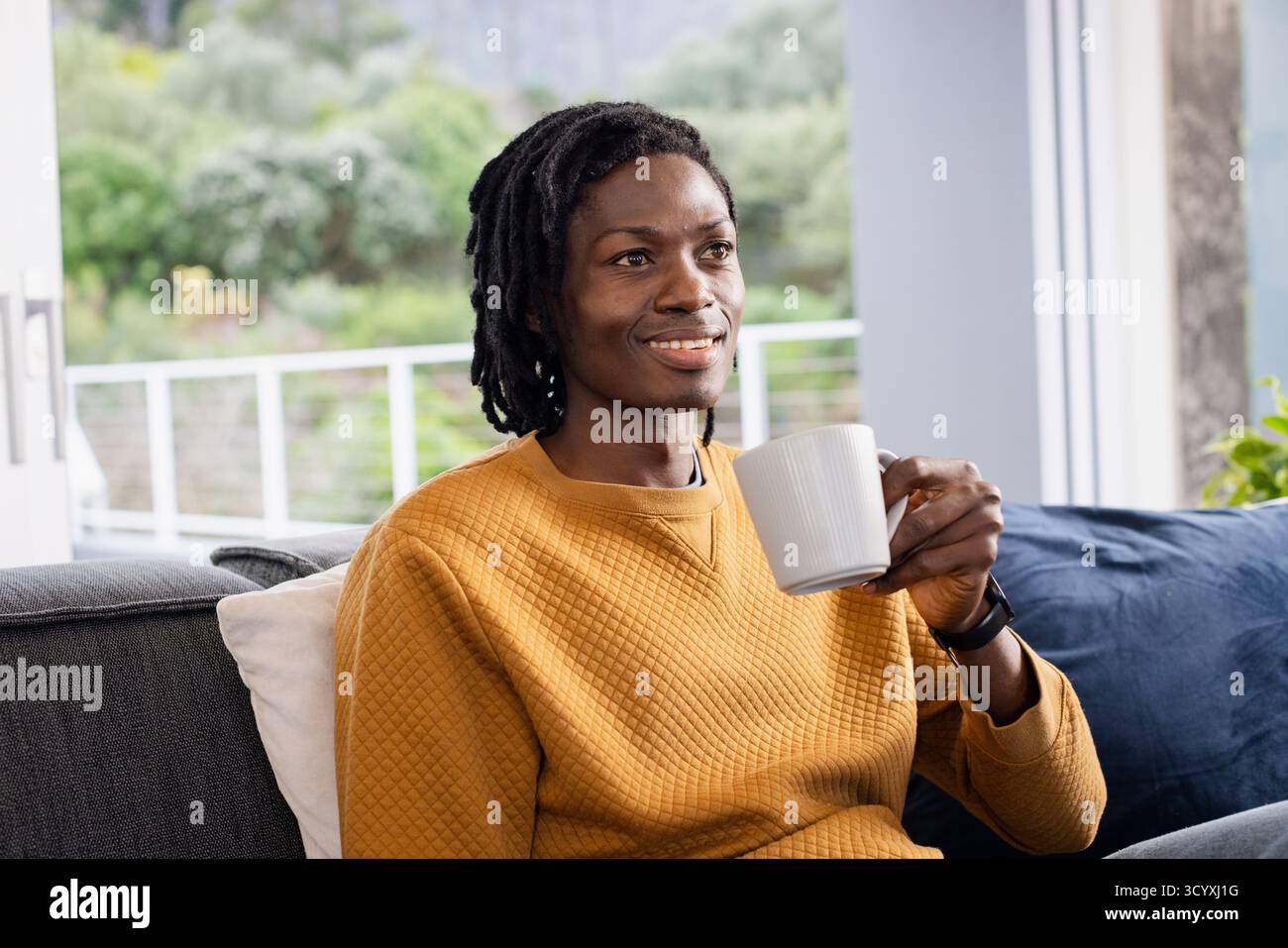 Afroamerikanischer Mann in Senfpullover, der zu Hause auf dem Sofa sitzt und einen Becher hält, der Smartwatch überprüft Stockfoto