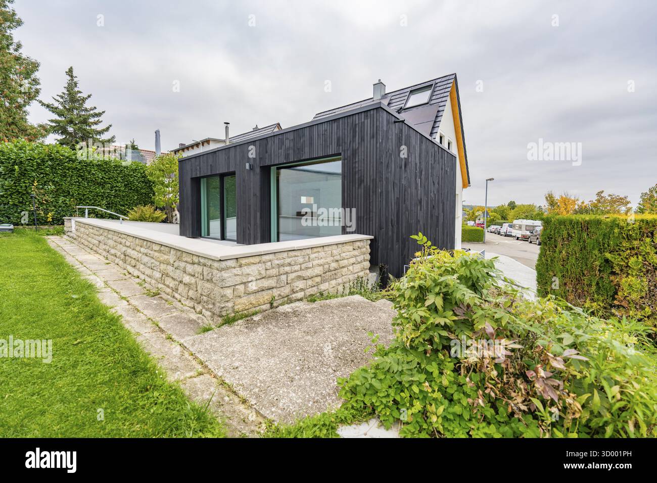 Schwarzer, modern gekleideter Anbau mit großen Glasflächen auf Rasen und Steinmauer in ruhiger Vorstadtlage mit bewölktem Himmel, Hausrenovierung, Klimaanlage Stockfoto