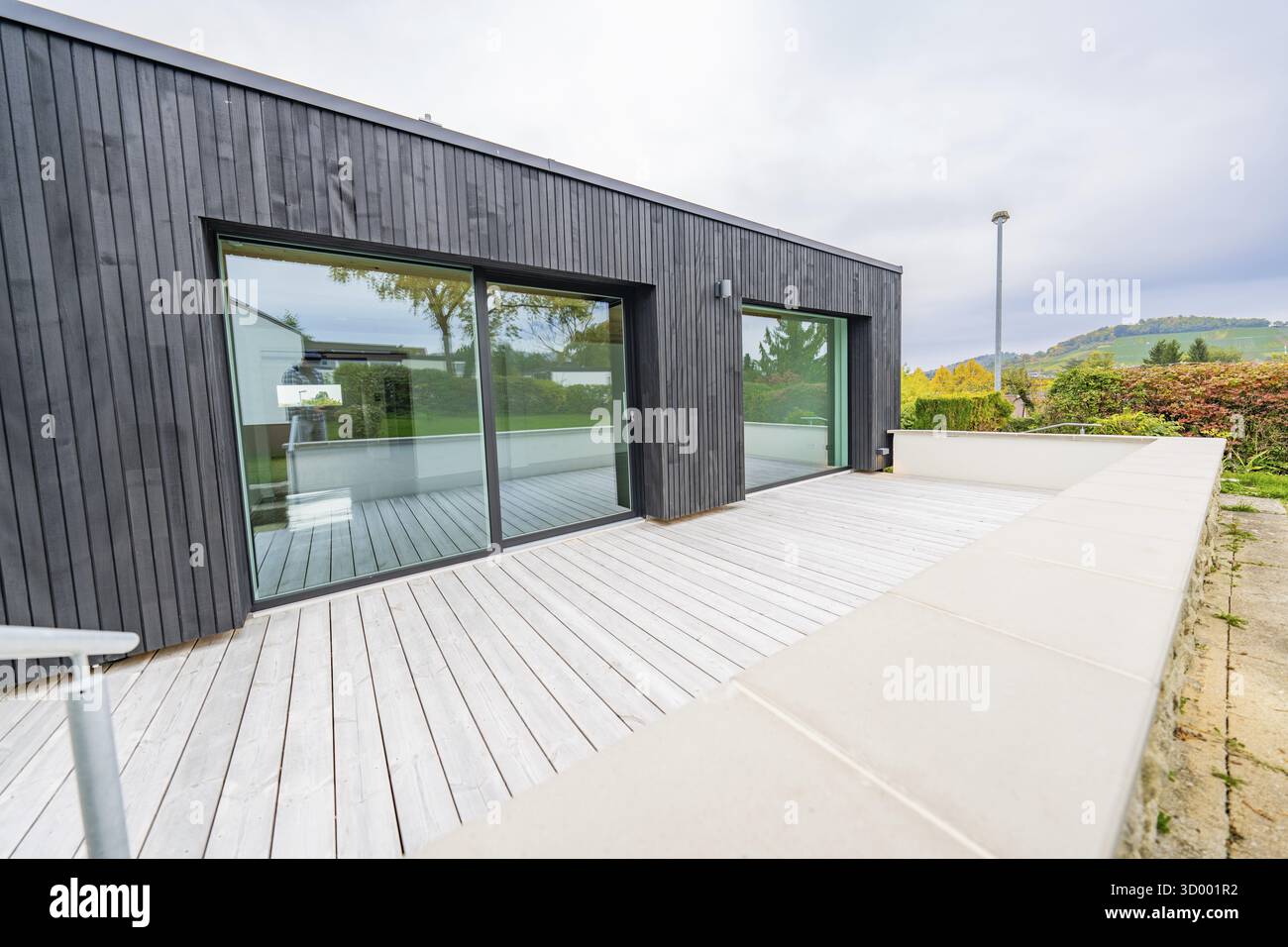 Große Terrasse mit heller Holzterrasse gegen schwarze Fassade und Schiebetüren, großer Blick auf die herbstliche Umgebung, Hausrenovierung, Klimaanlage Stockfoto