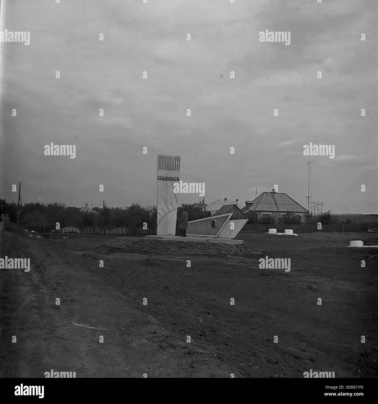 Dieses Archivfoto aus dem Jahr 1966 von Sloviansk, Ukrainische SSR, zeigt das Haupteingangsschild der Stadt oder die Stele. Dieses Denkmal ist ein perfektes Beispiel für die sowjetische Moderne, die in den 1960er Jahren populär war Das senkrechte Element, das einem Sonnenstrahl oder einem Segel ähnelt, trägt den Namen der Stadt „СЛАВЯНСК“ (SLAWJANSK) in kyrillischer Schrift. Die gesamte Struktur hat dynamische, kantige Formen. Es liegt an einer unbefestigten Straße am Stadtrand, mit einfachen Landhäusern im Hintergrund. Dieses Wahrzeichen war ein Symbol des Stolzes der Bürger, ein wichtiger Bestandteil des friedlichen Donbass Stockfoto